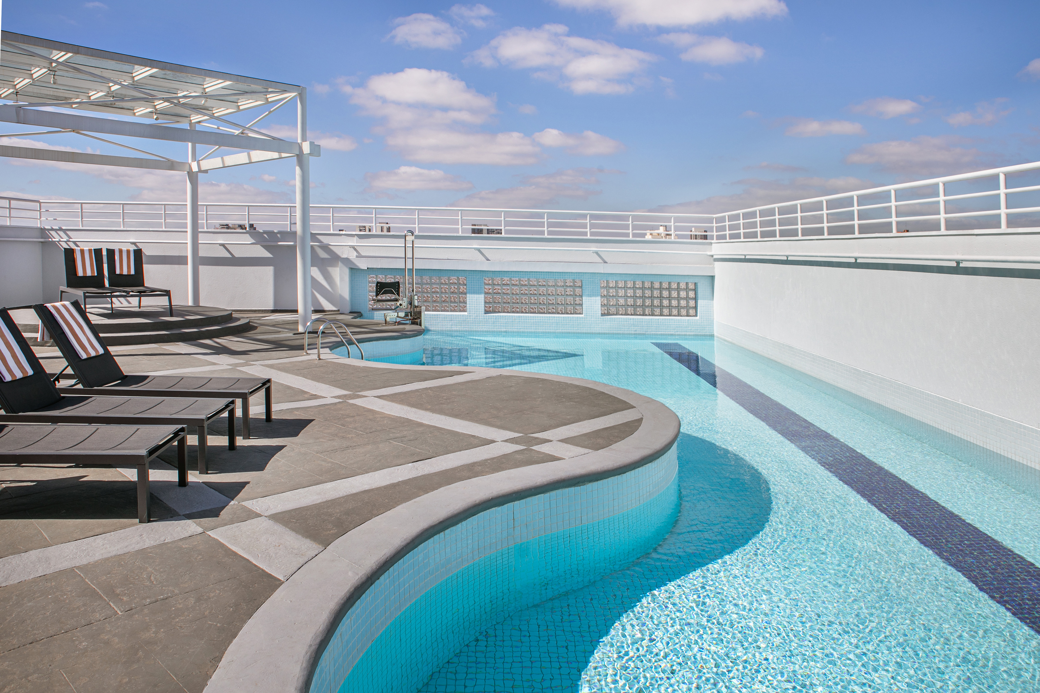Rooftop swimming pool with modern lounge chairs and white pergola under blue sky.