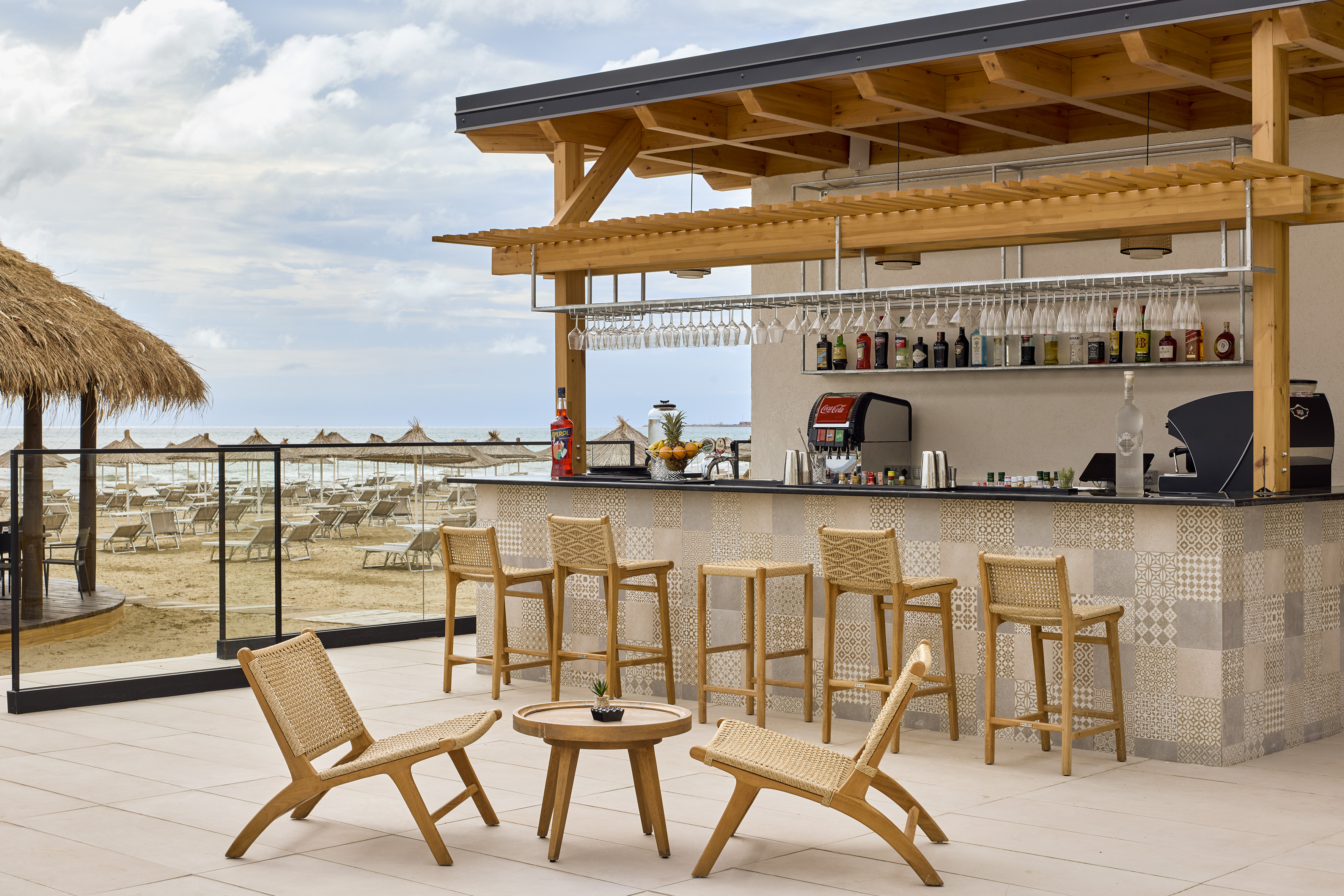 Beach bar with wooden structure, patterned counter, seating, and beach view with umbrellas.