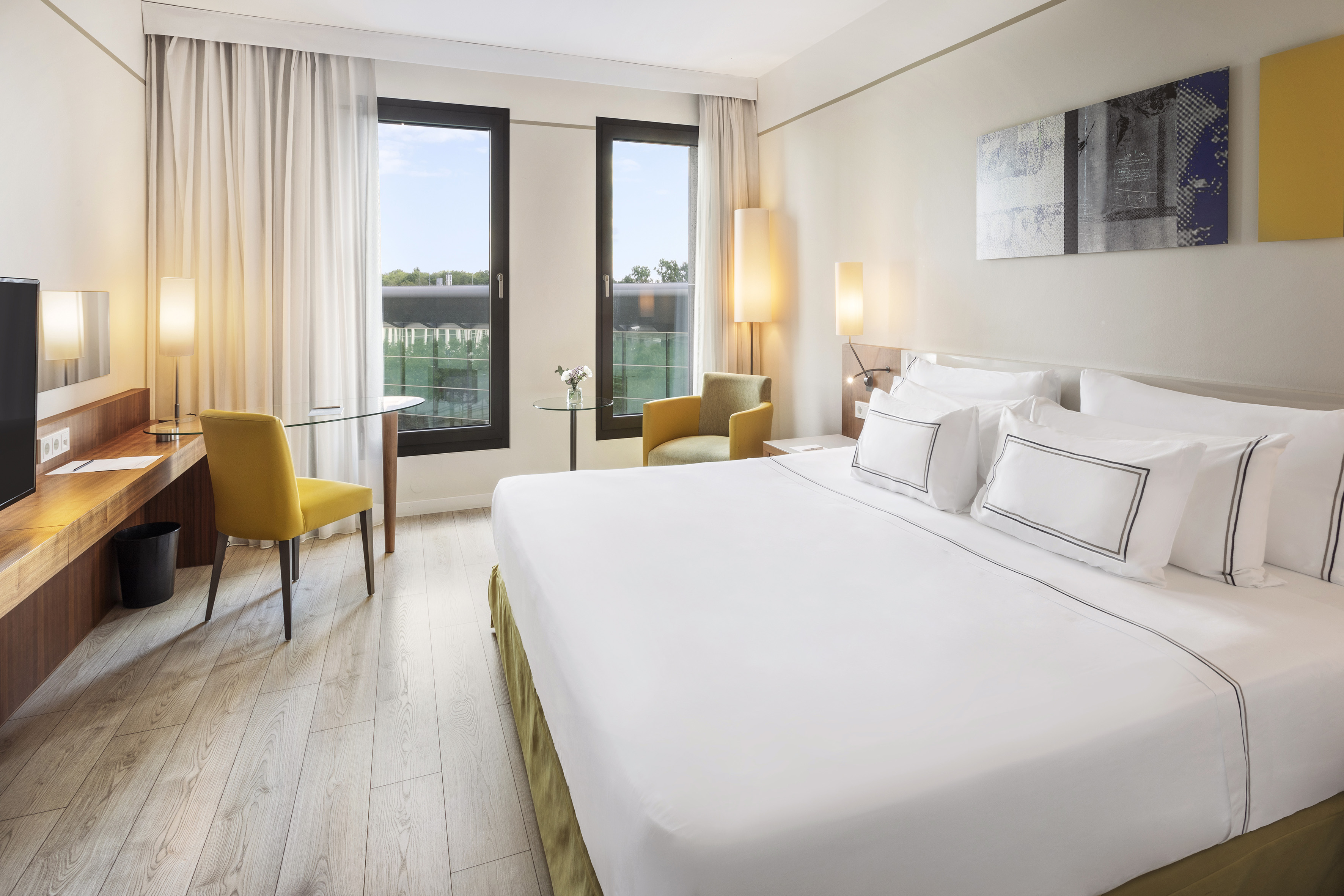 Modern hotel room with king bed, desk, armchair, and bright windows.