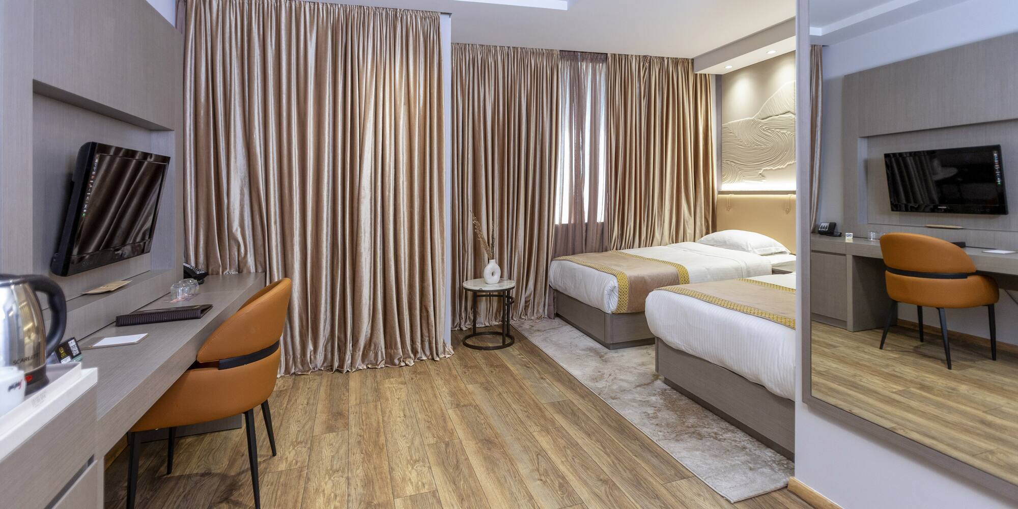 Modern hotel twin room with wood floor, desk, TV, and beige curtains.