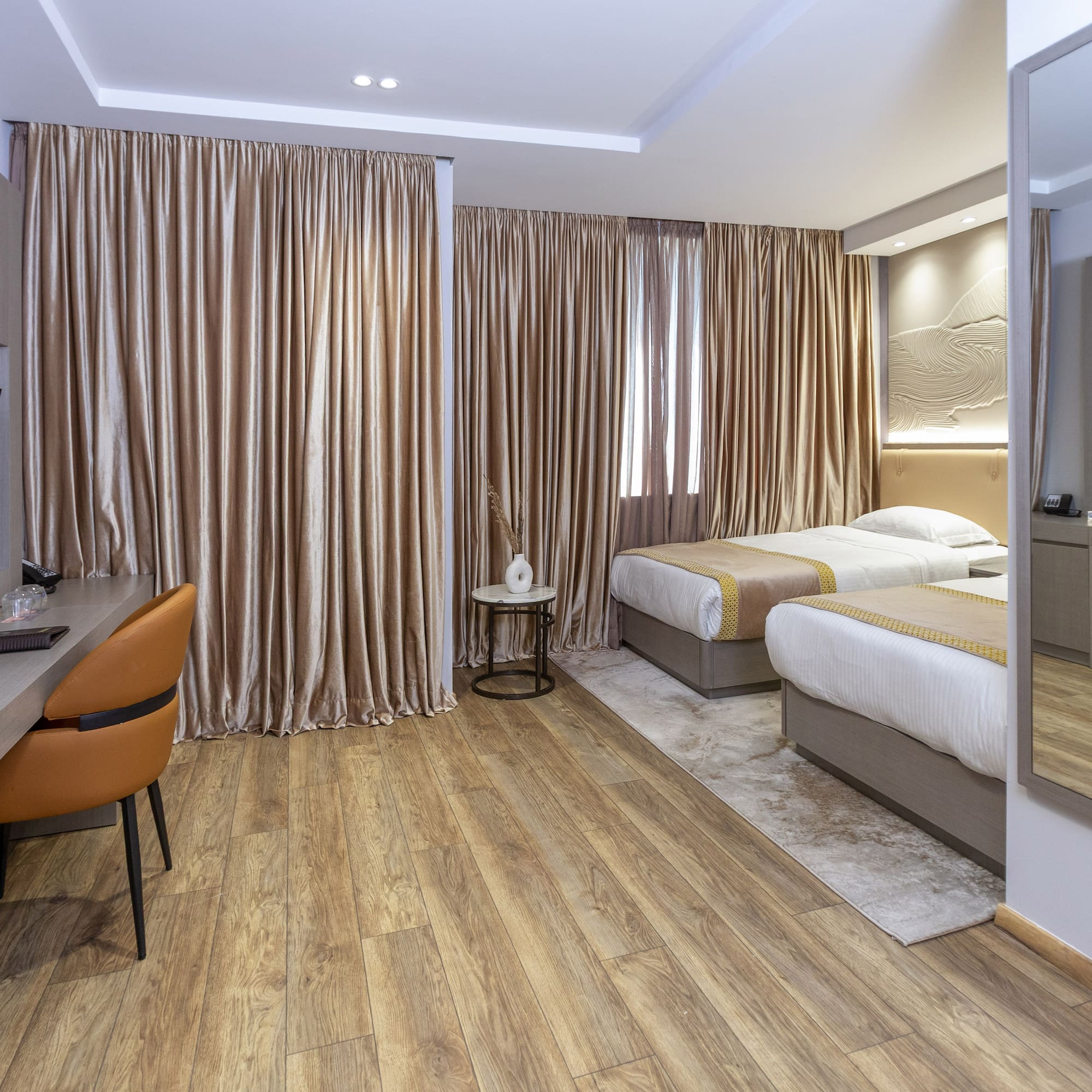 Modern hotel twin room with wood floor, desk, TV, and beige curtains.