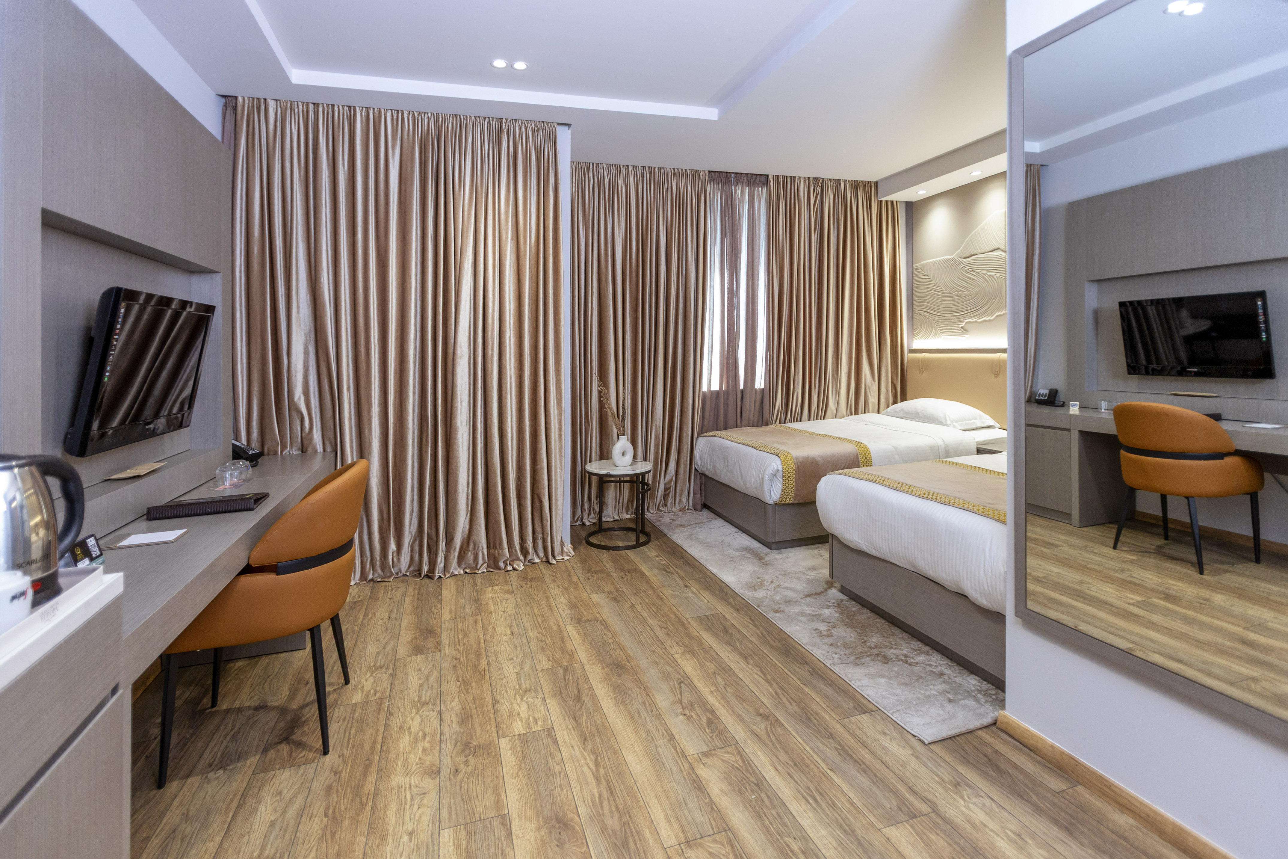 Modern hotel twin room with wood floor, desk, TV, and beige curtains.