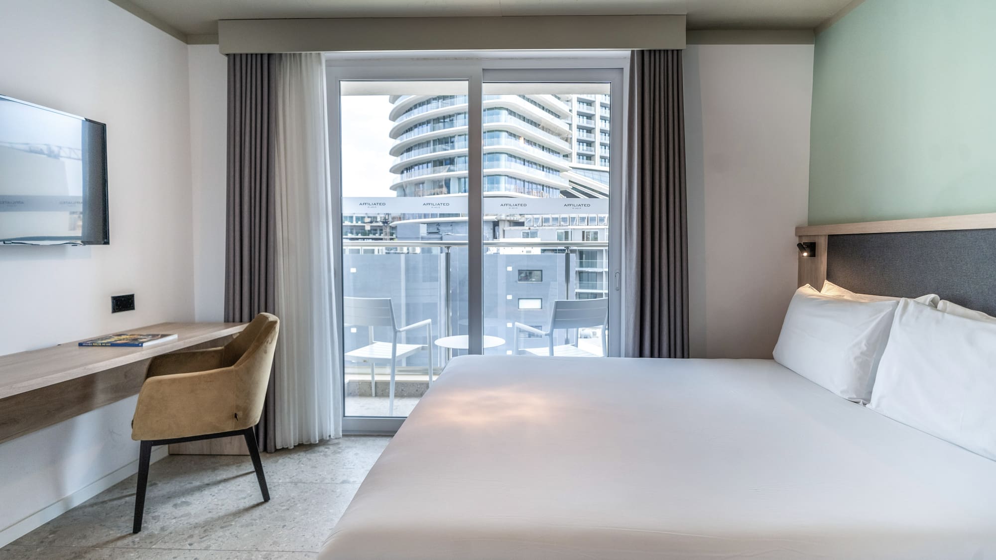 Modern hotel room with bed, desk, and balcony offering city views.