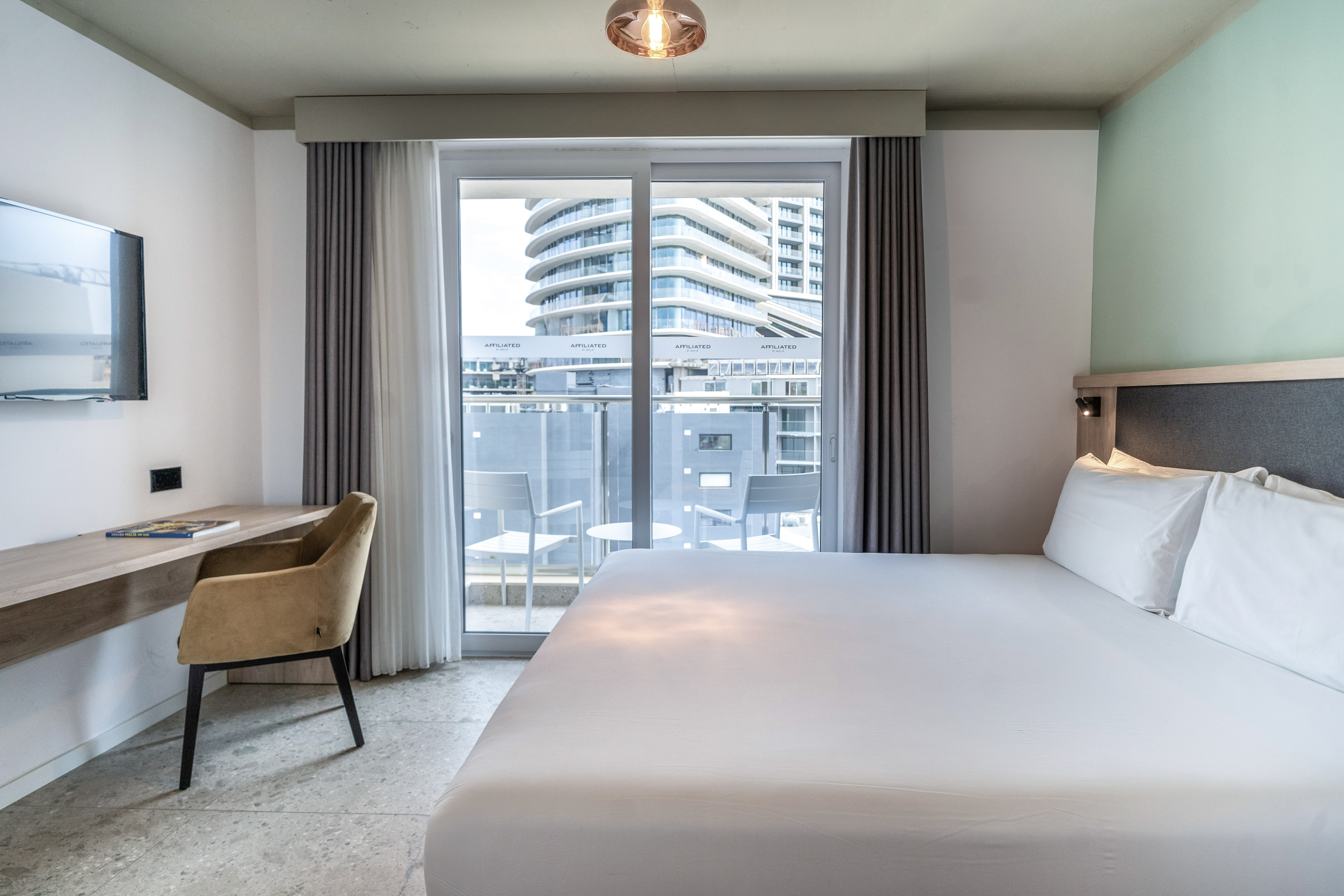 Modern hotel room with bed, desk, and balcony offering city views.