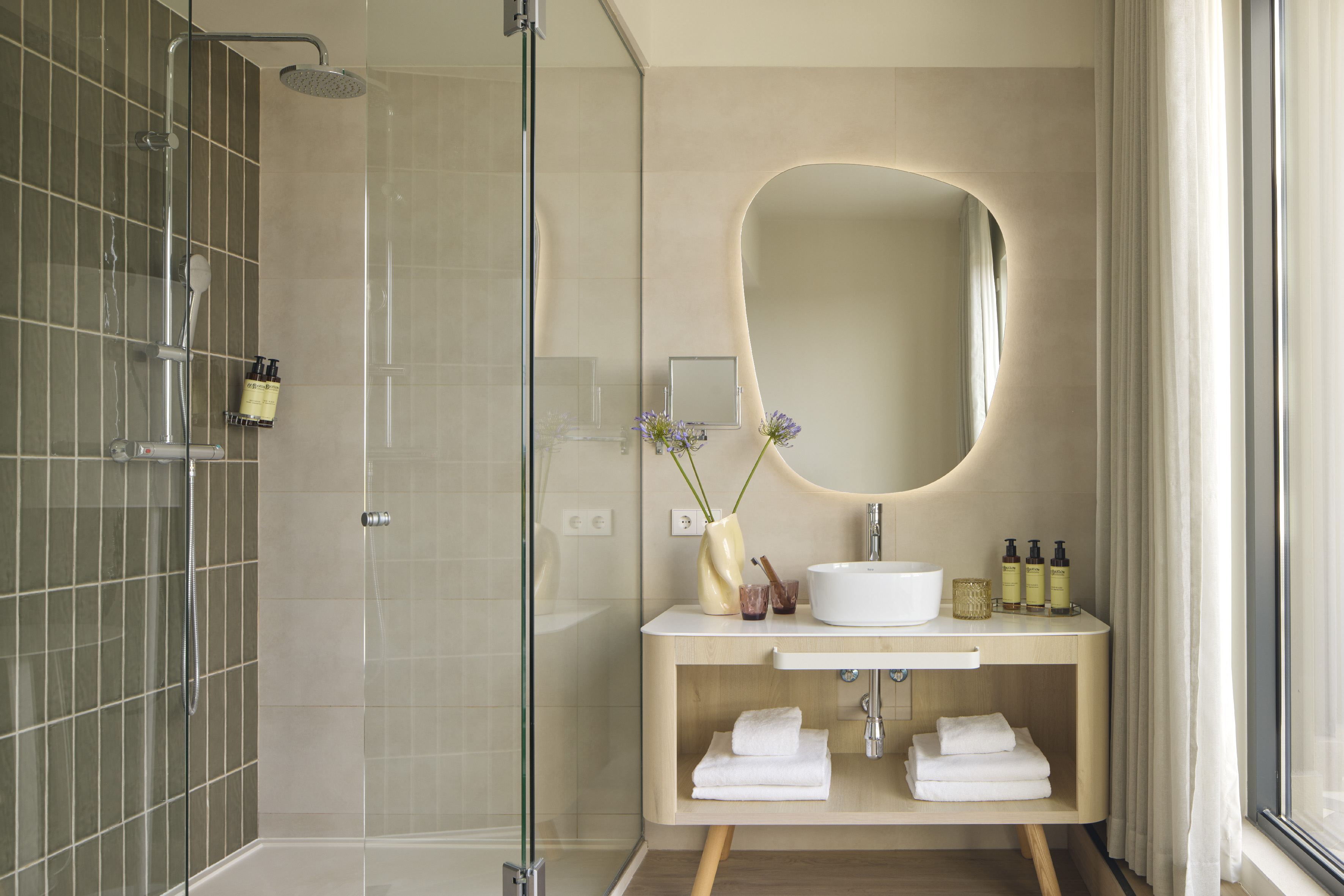 Modern bathroom with glass shower, illuminated mirror, wood vanity, and white sink.