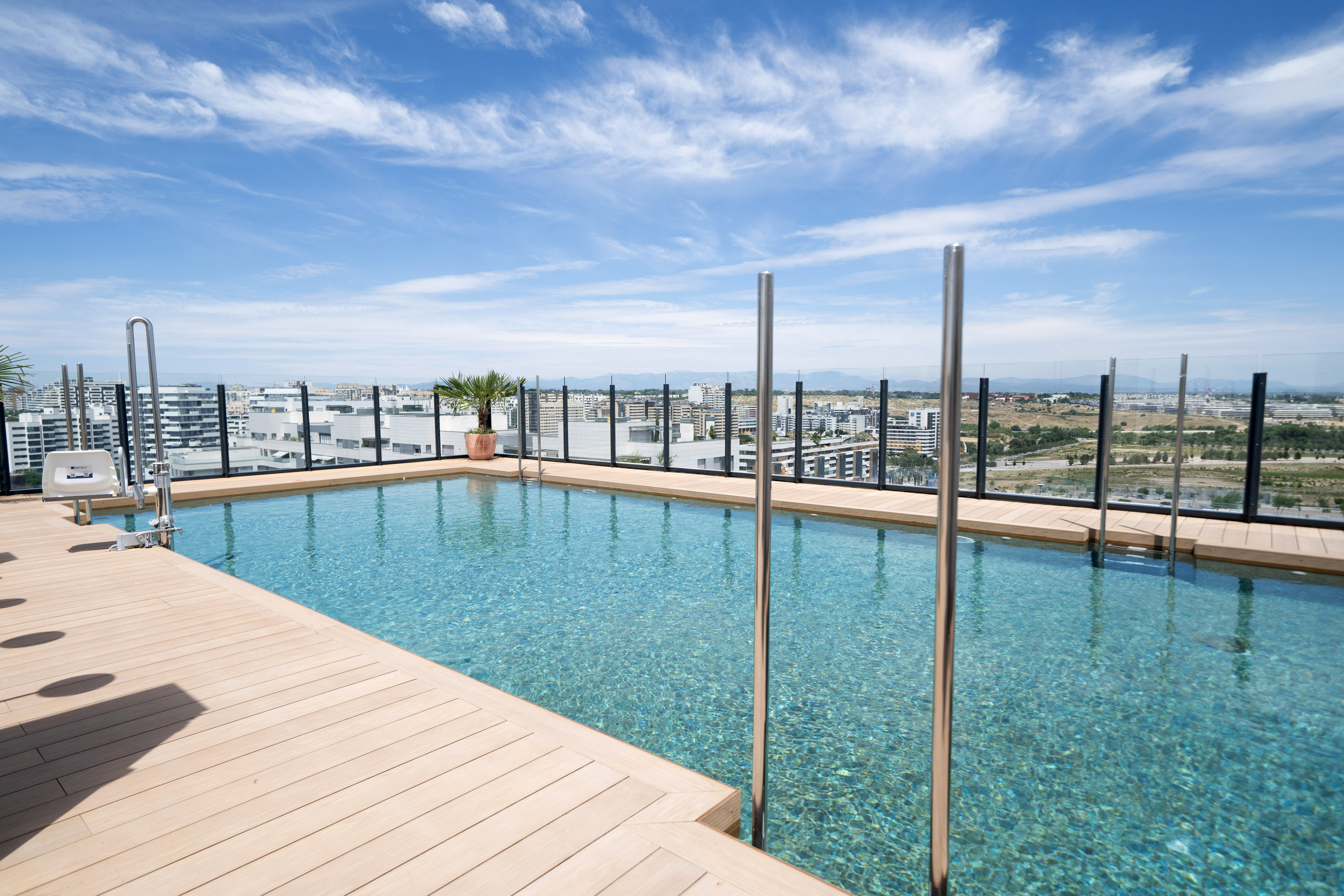 Accessible rooftop pool with wooden deck overlooking a city view and bright sky.