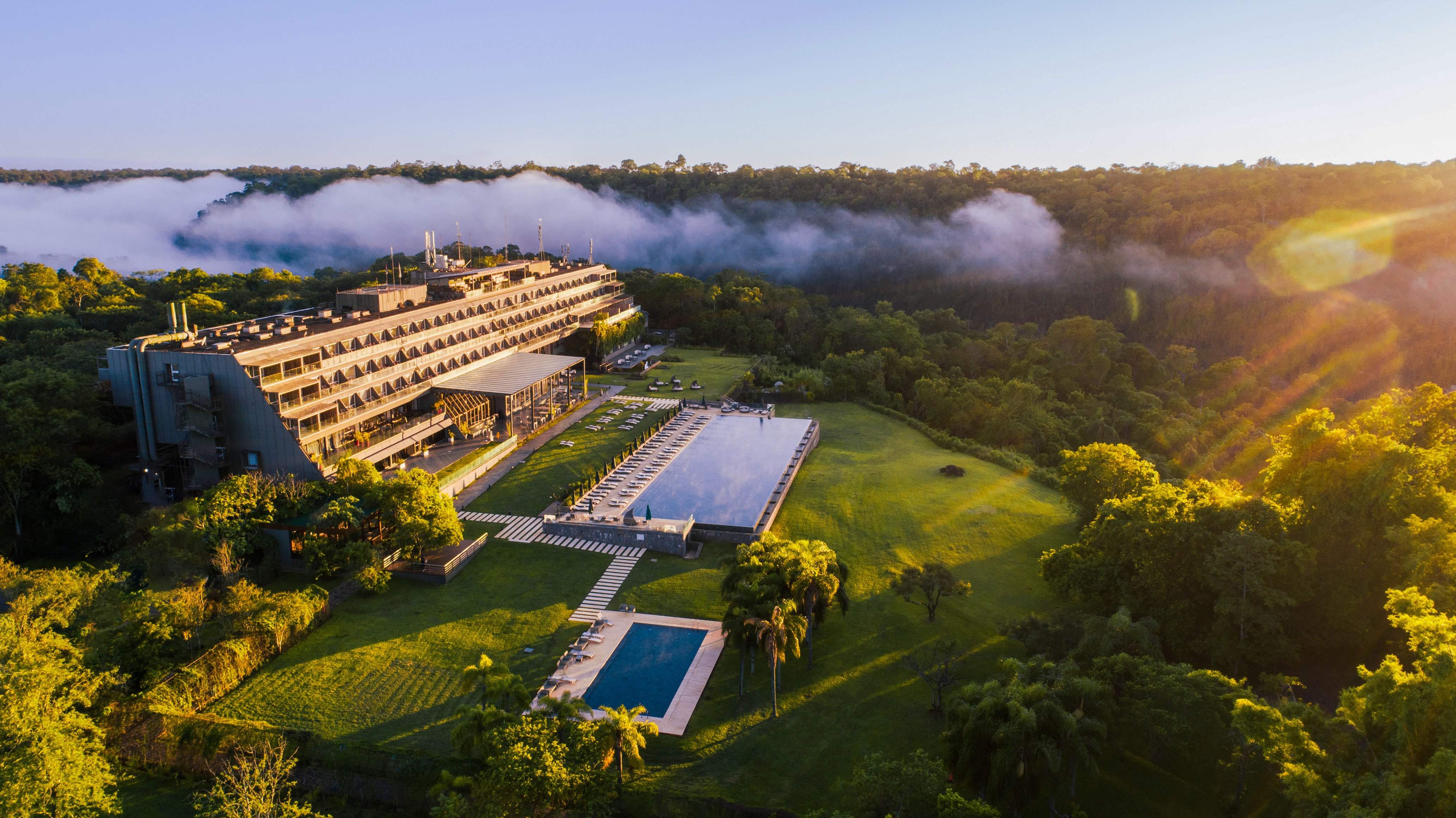 Aerial view: Luxury hotel with two pools, lush jungle, and mist at golden hour.