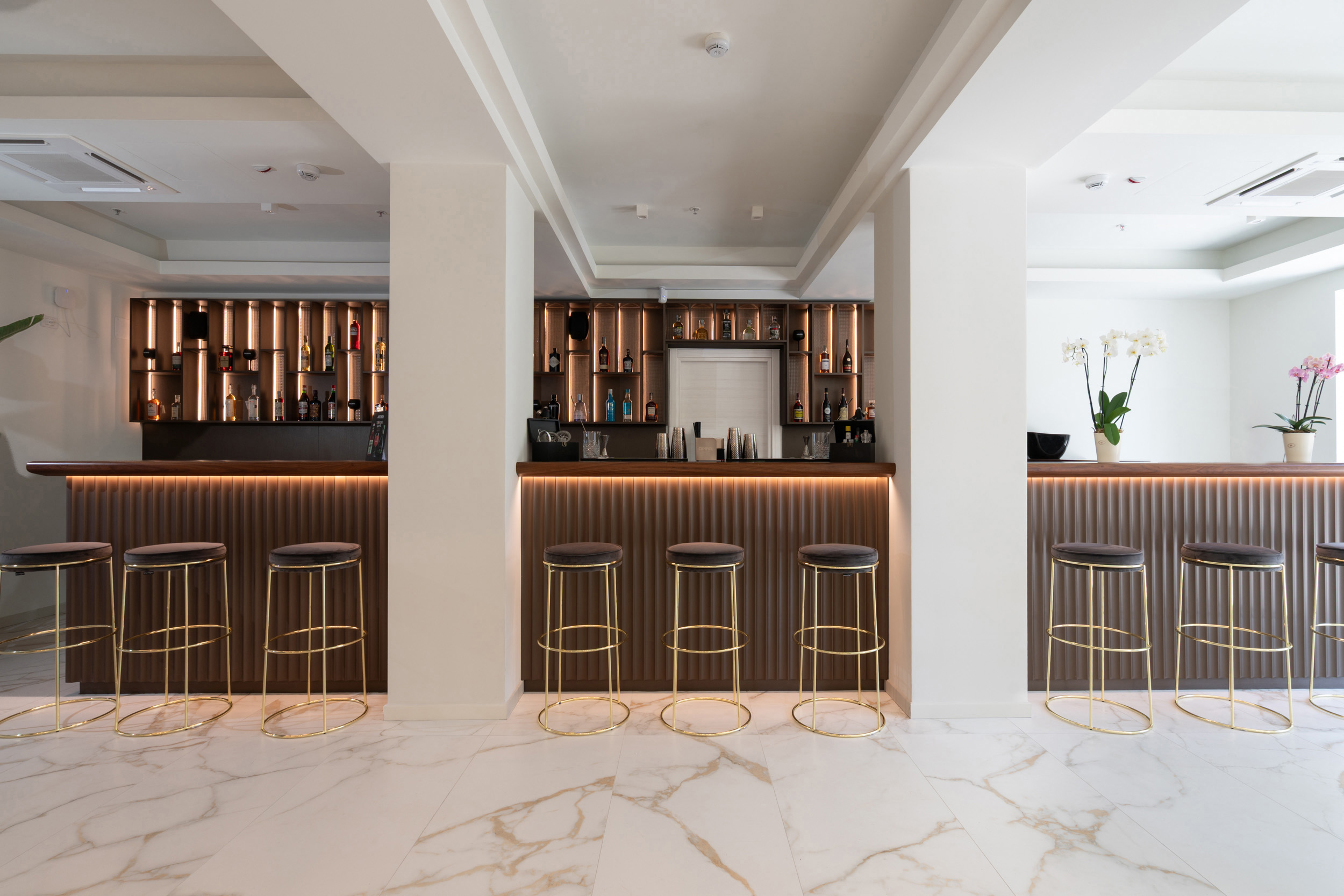 Modern hotel bar with backlit liquor shelves, gold bar stools, and marble floor.