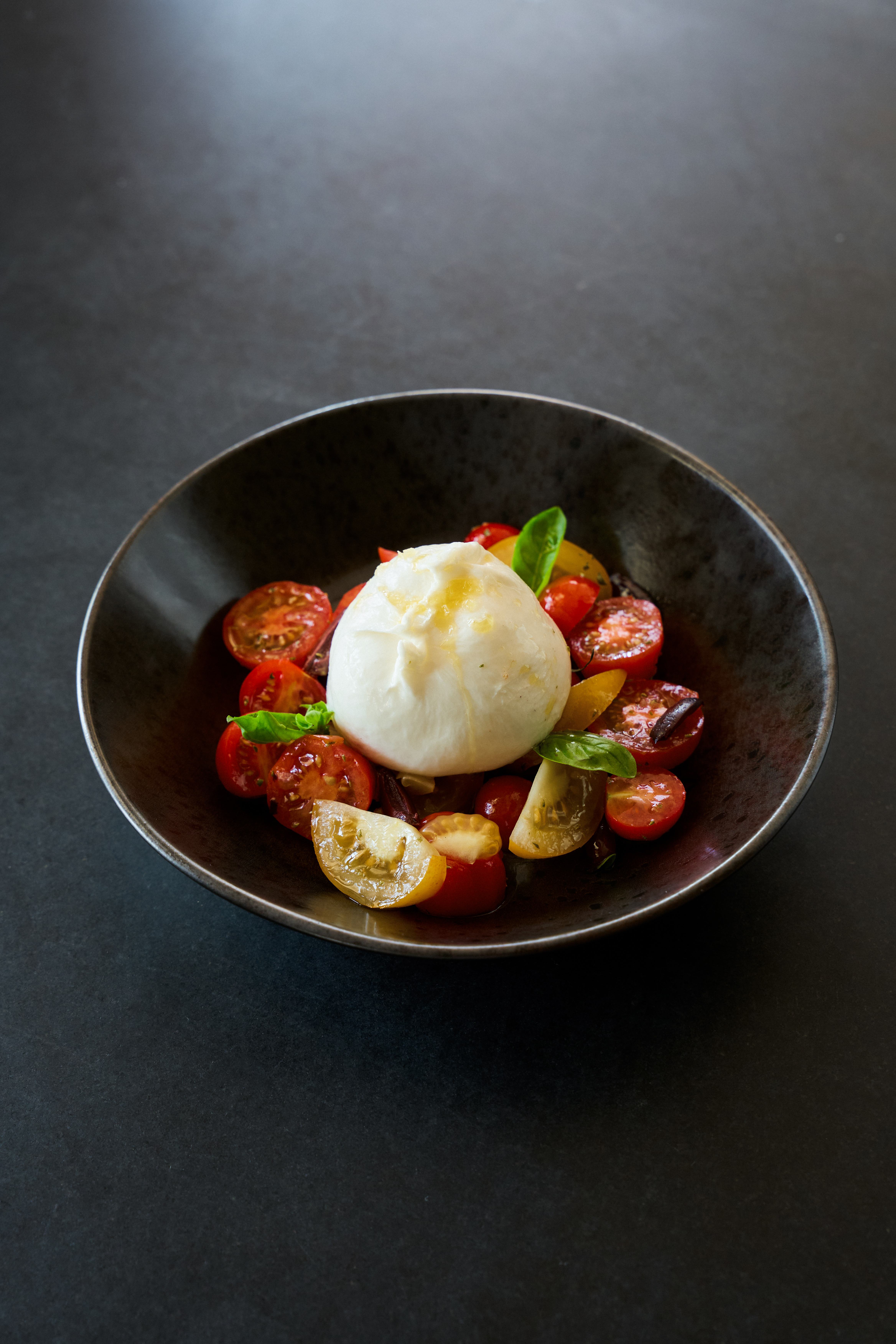 Burrata cheese with red and yellow cherry tomatoes and fresh basil in a dark bowl.