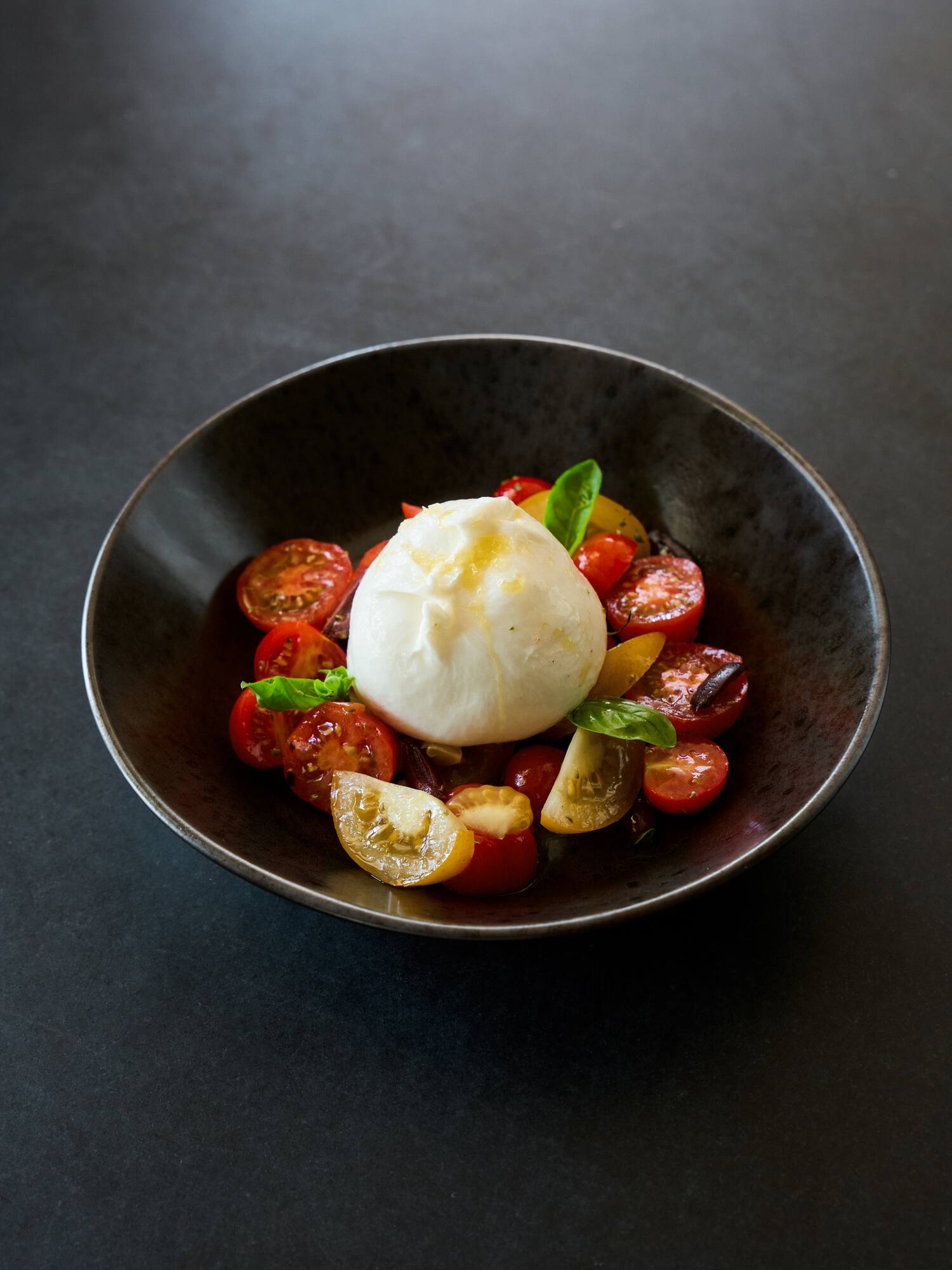Burrata cheese with red and yellow cherry tomatoes and fresh basil in a dark bowl.