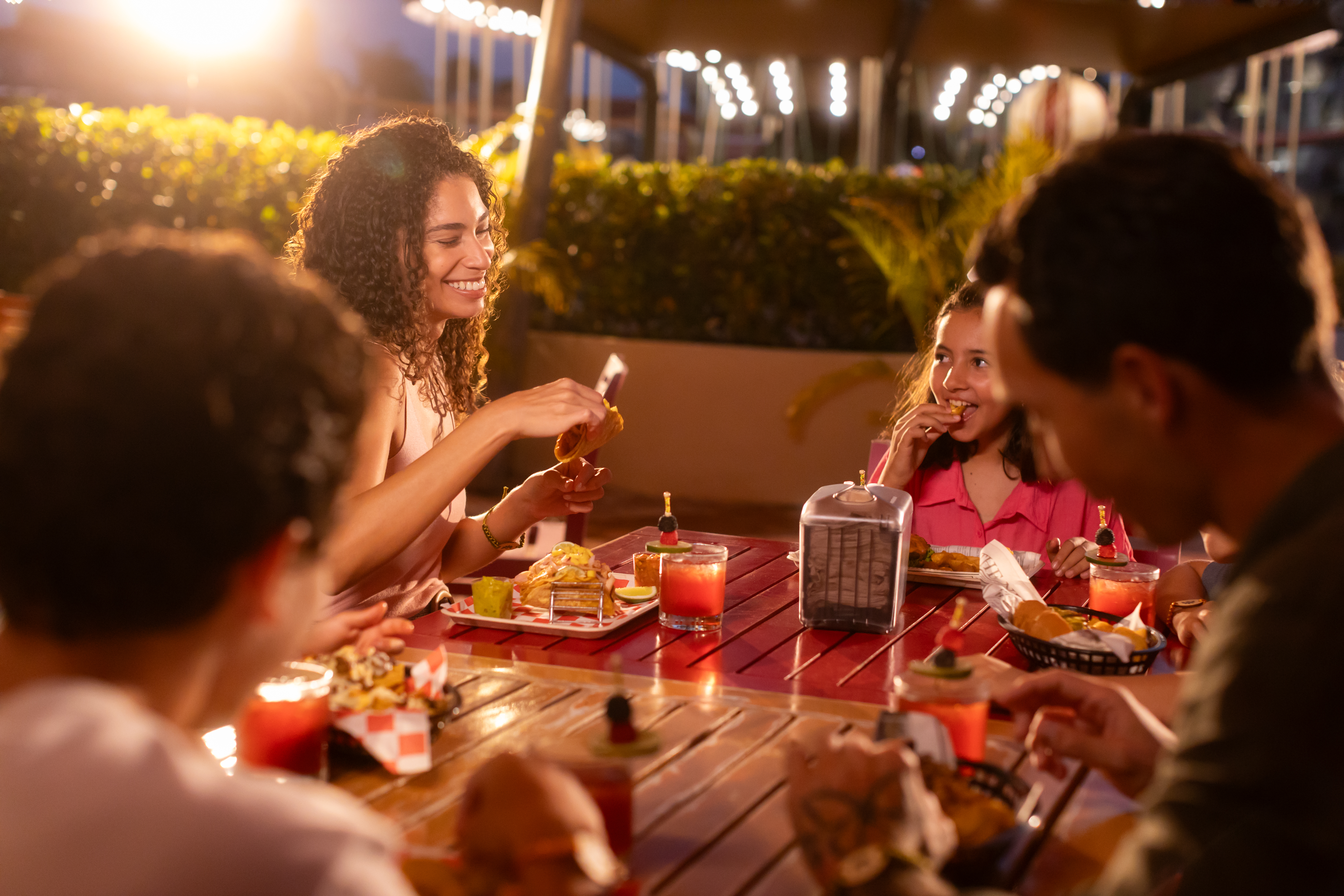 Happy family dining outdoors, eating tacos and drinks at a brightly lit restaurant.
