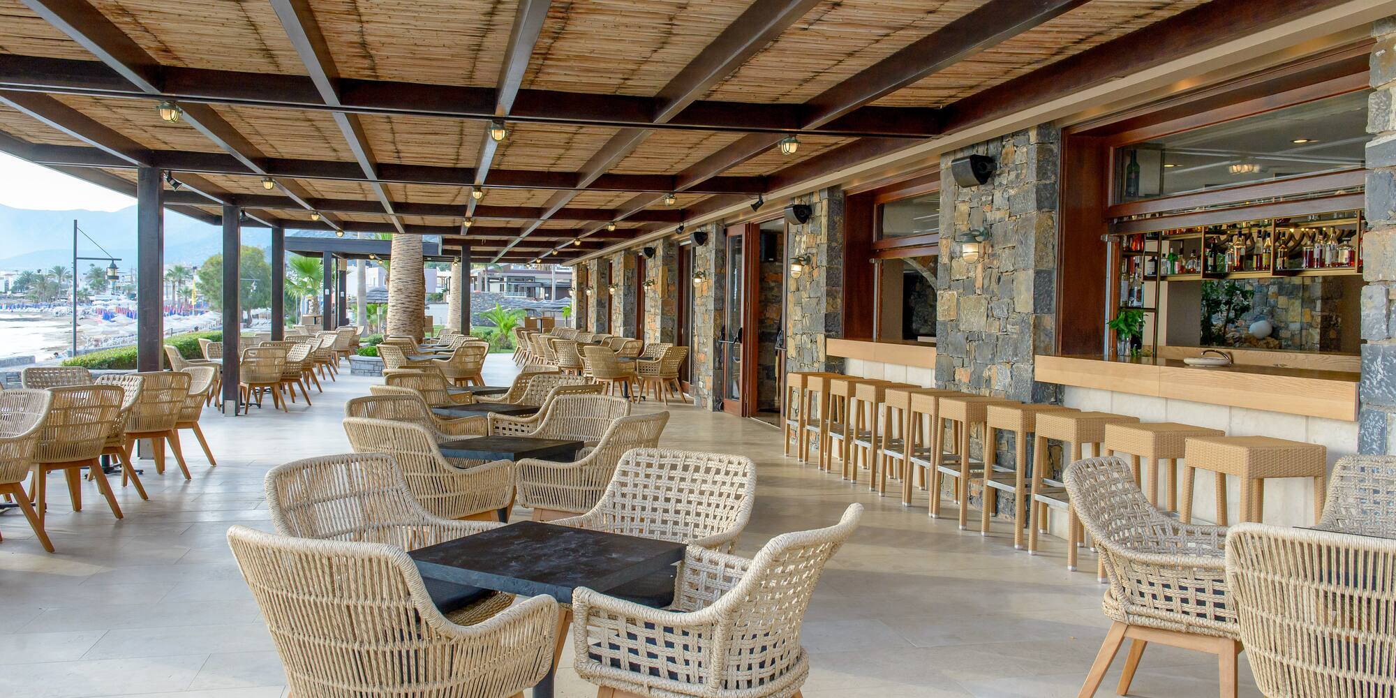 Outdoor restaurant terrace with woven wicker chairs, stone bar, and beach view.