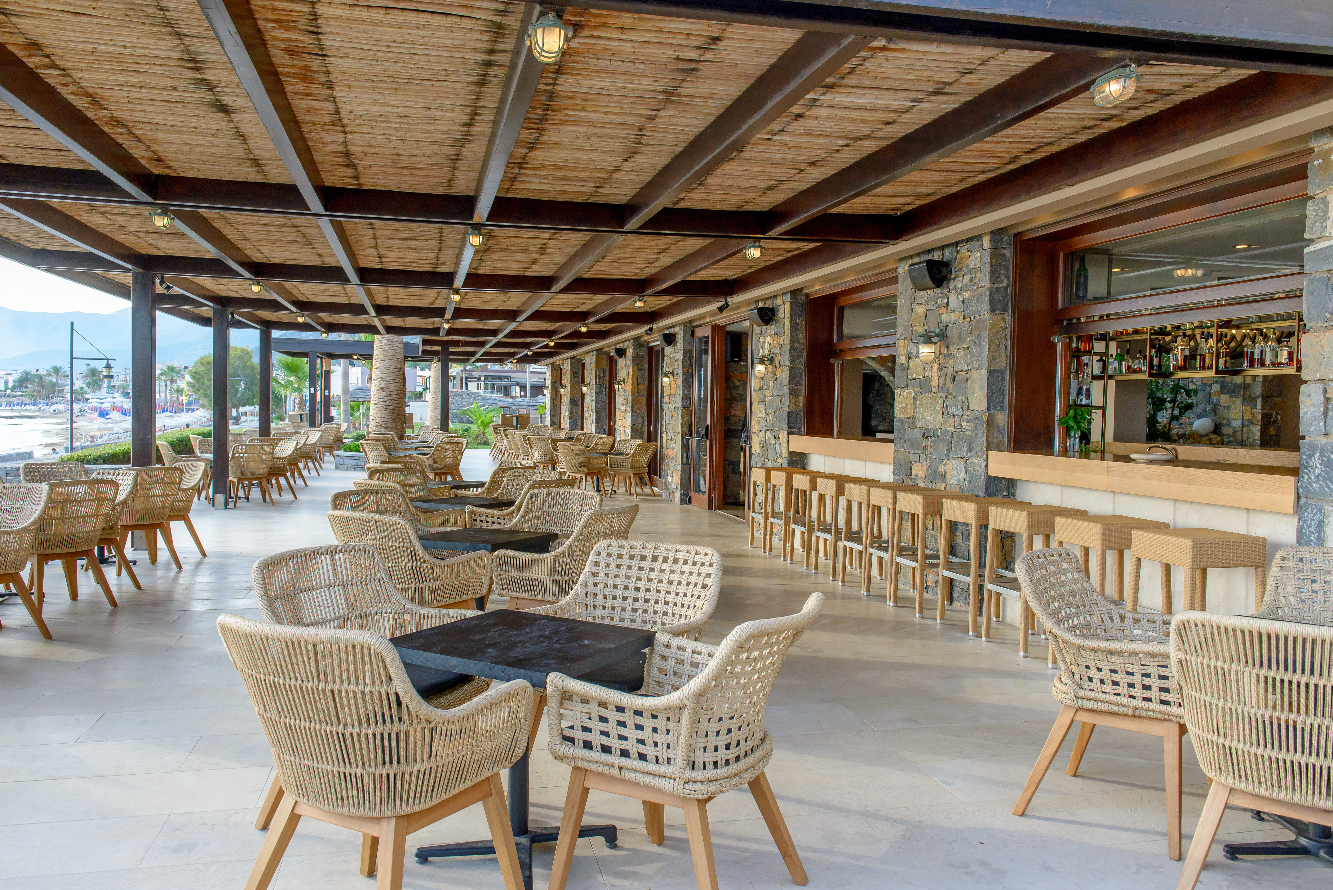 Outdoor restaurant terrace with woven wicker chairs, stone bar, and beach view.