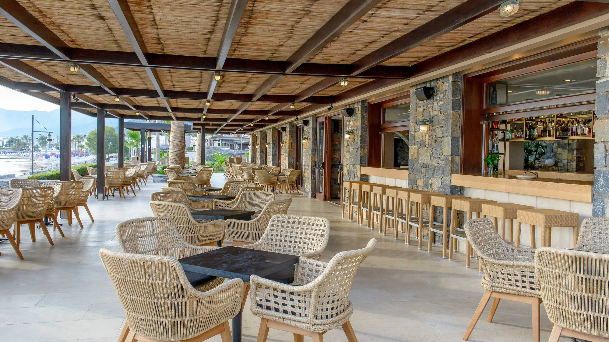 Outdoor restaurant terrace with woven wicker chairs, stone bar, and beach view.