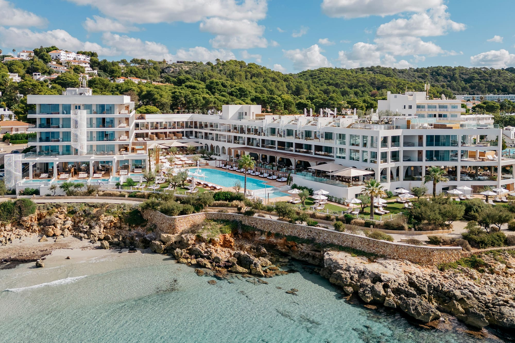 Aerial view of modern resort, clear blue sea, rocky cove, and large pool.