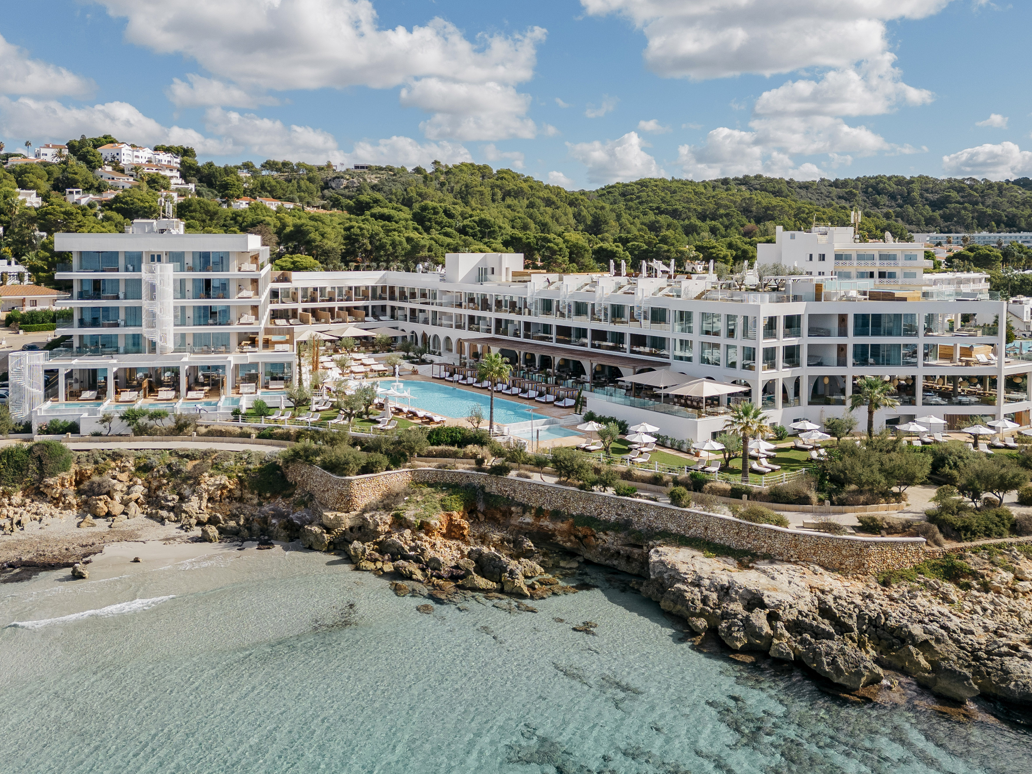 Aerial view of modern resort, clear blue sea, rocky cove, and large pool.