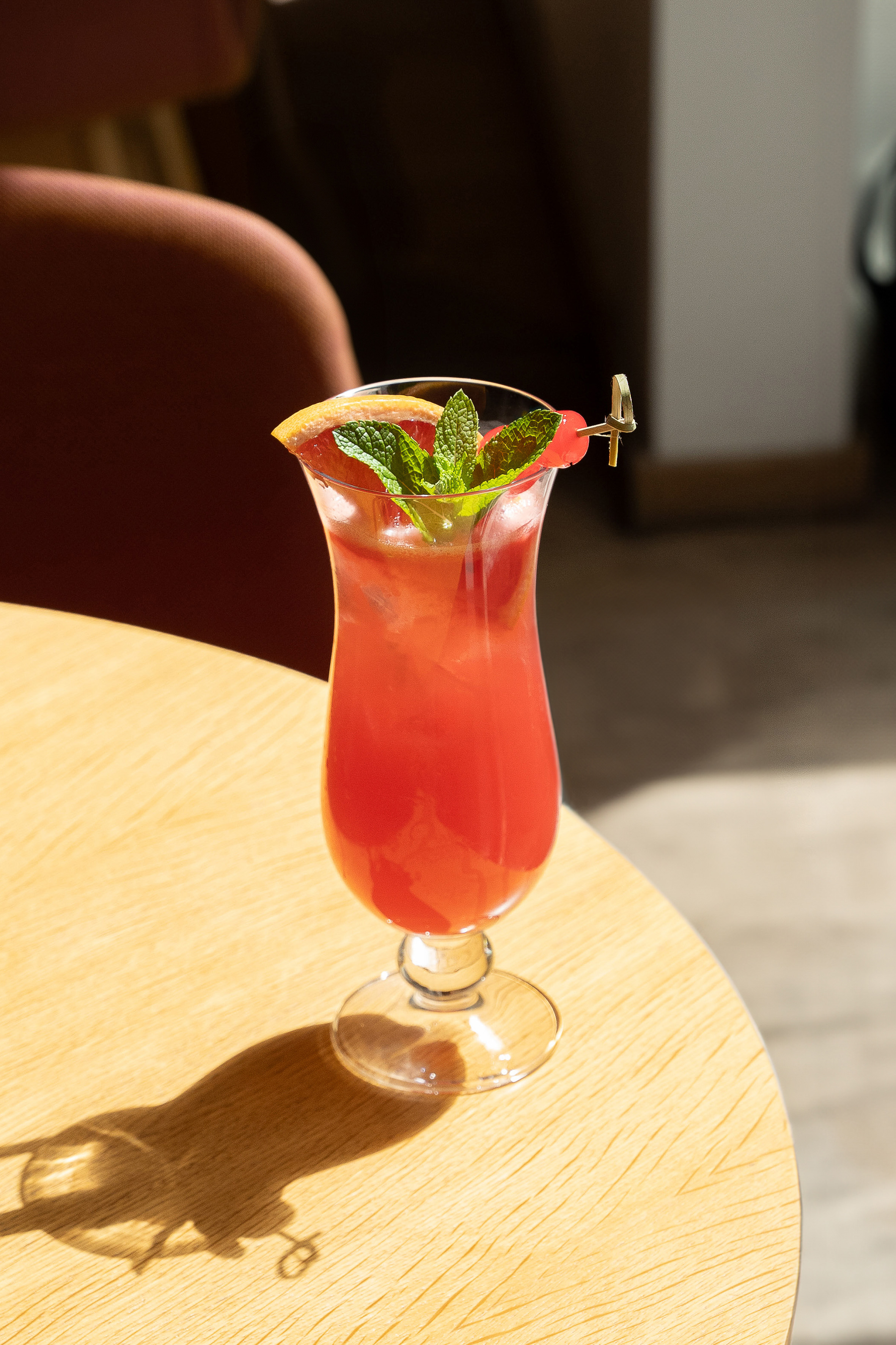 Red cocktail in tall glass with grapefruit, mint, and cherry on wood table.