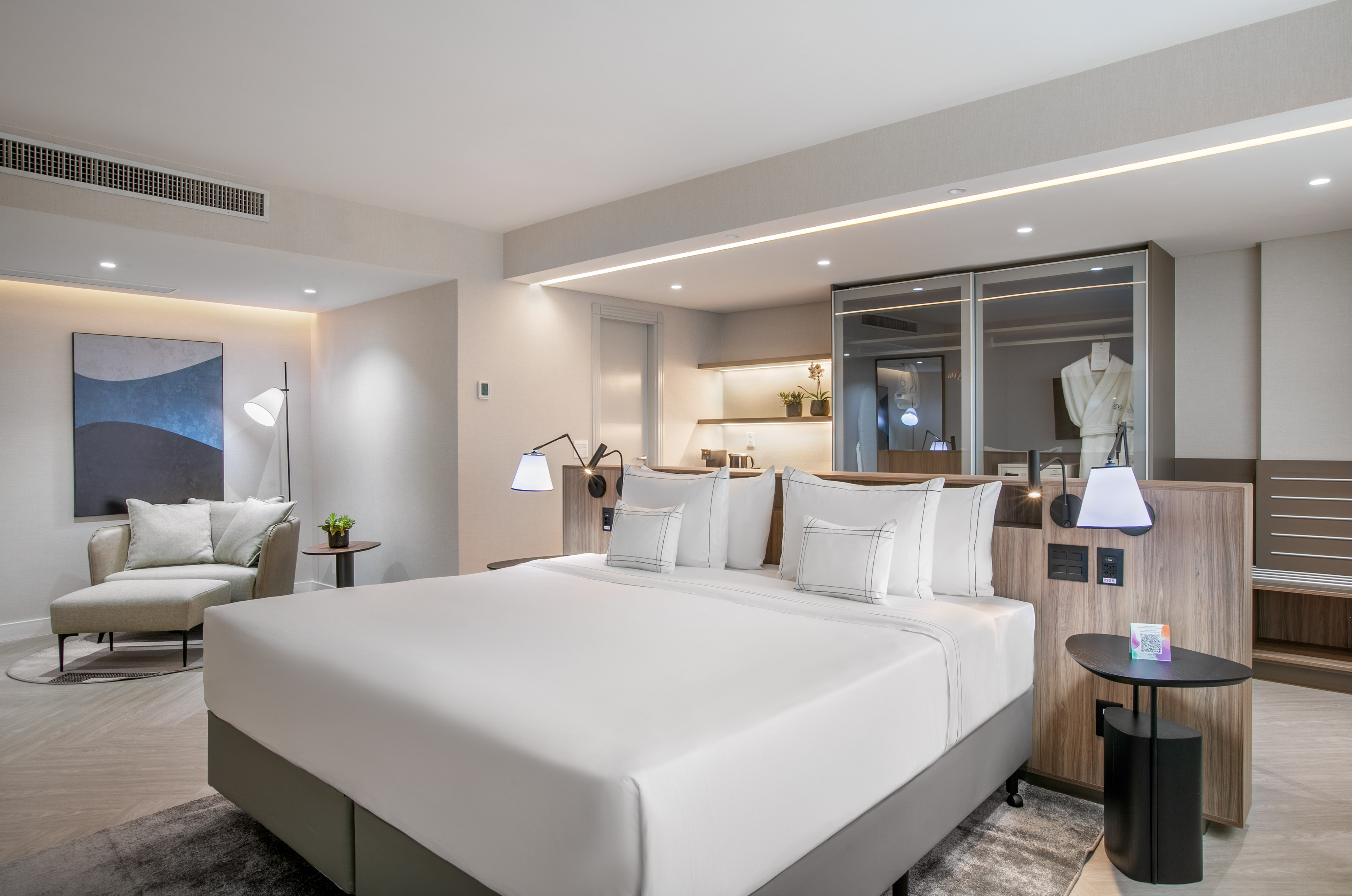 Modern hotel bedroom with large white bed, lounge chair, and built-in closet.