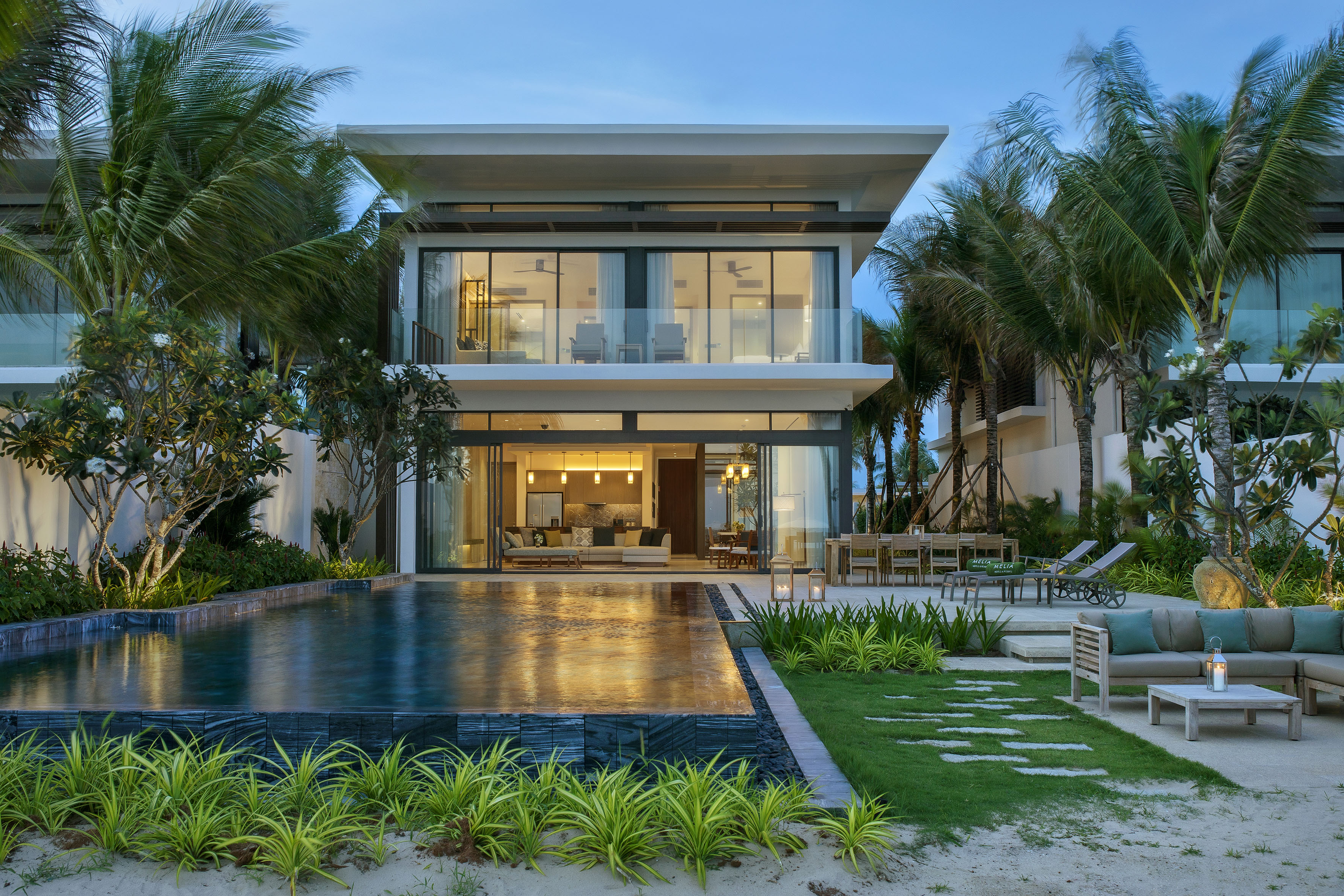 Luxury modern villa with infinity pool, palm trees, and outdoor seating at dusk.