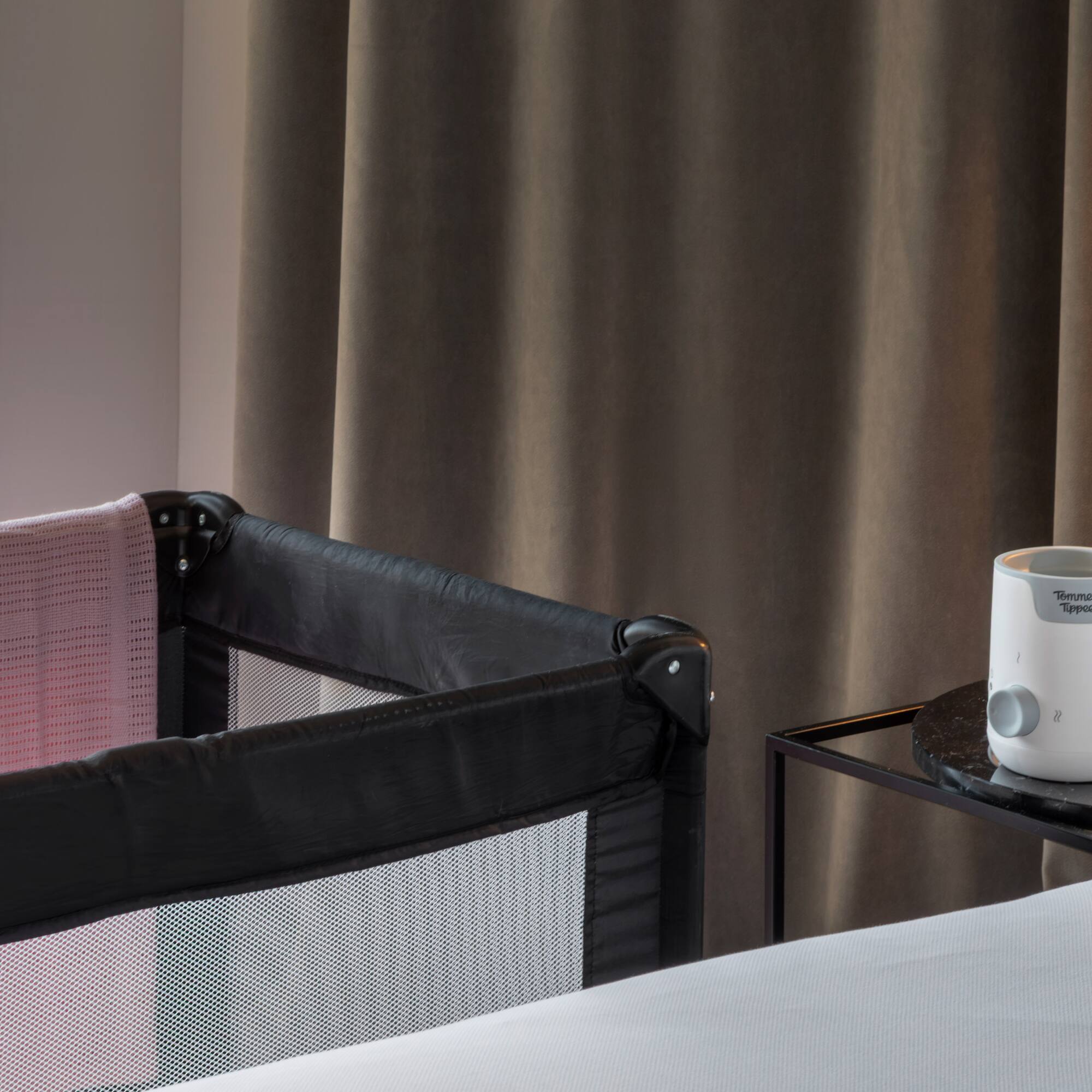 Travel crib with pink blanket and baby bottle warmer in hotel room.