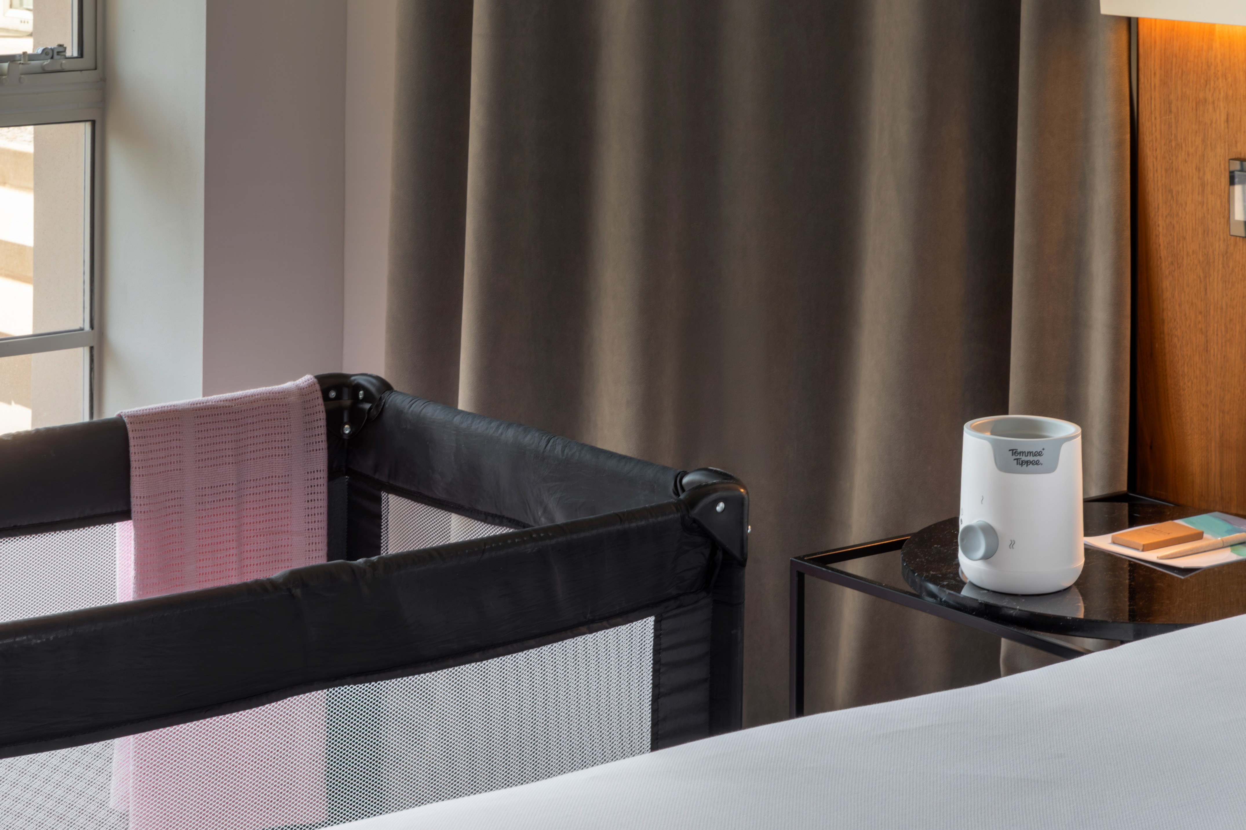 Travel crib with pink blanket and baby bottle warmer in hotel room.