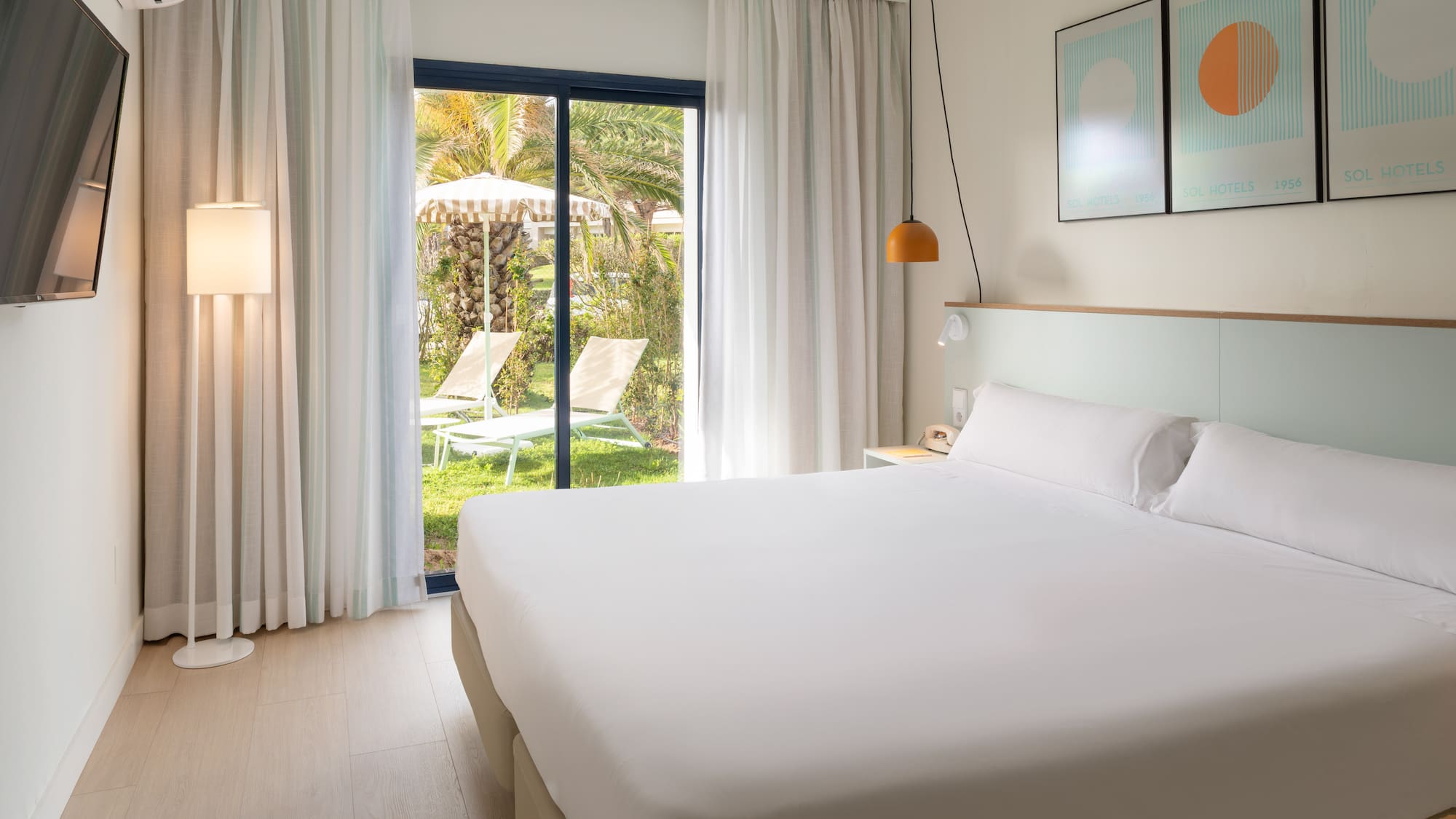 Bright hotel bedroom with white bed, glass door to garden patio, and sun loungers.