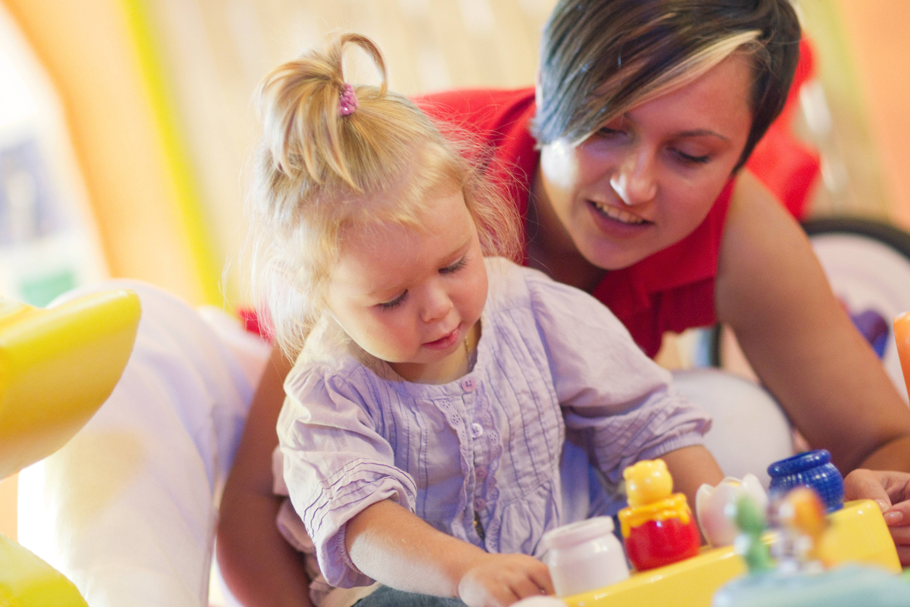 Adult woman smiles as toddler plays with colorful early learning toys.