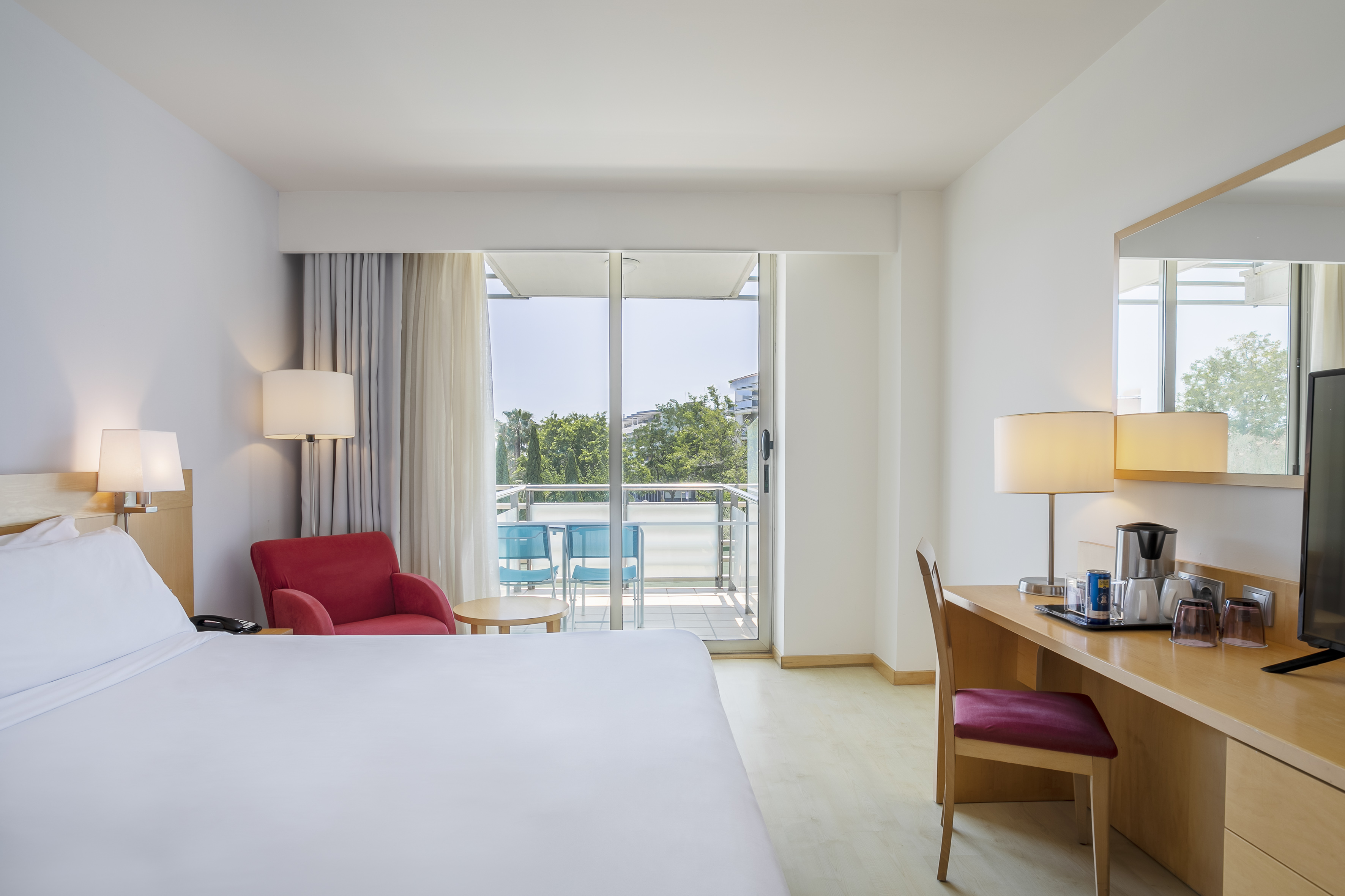 Bright hotel room with white bed, red armchair, desk, and balcony.