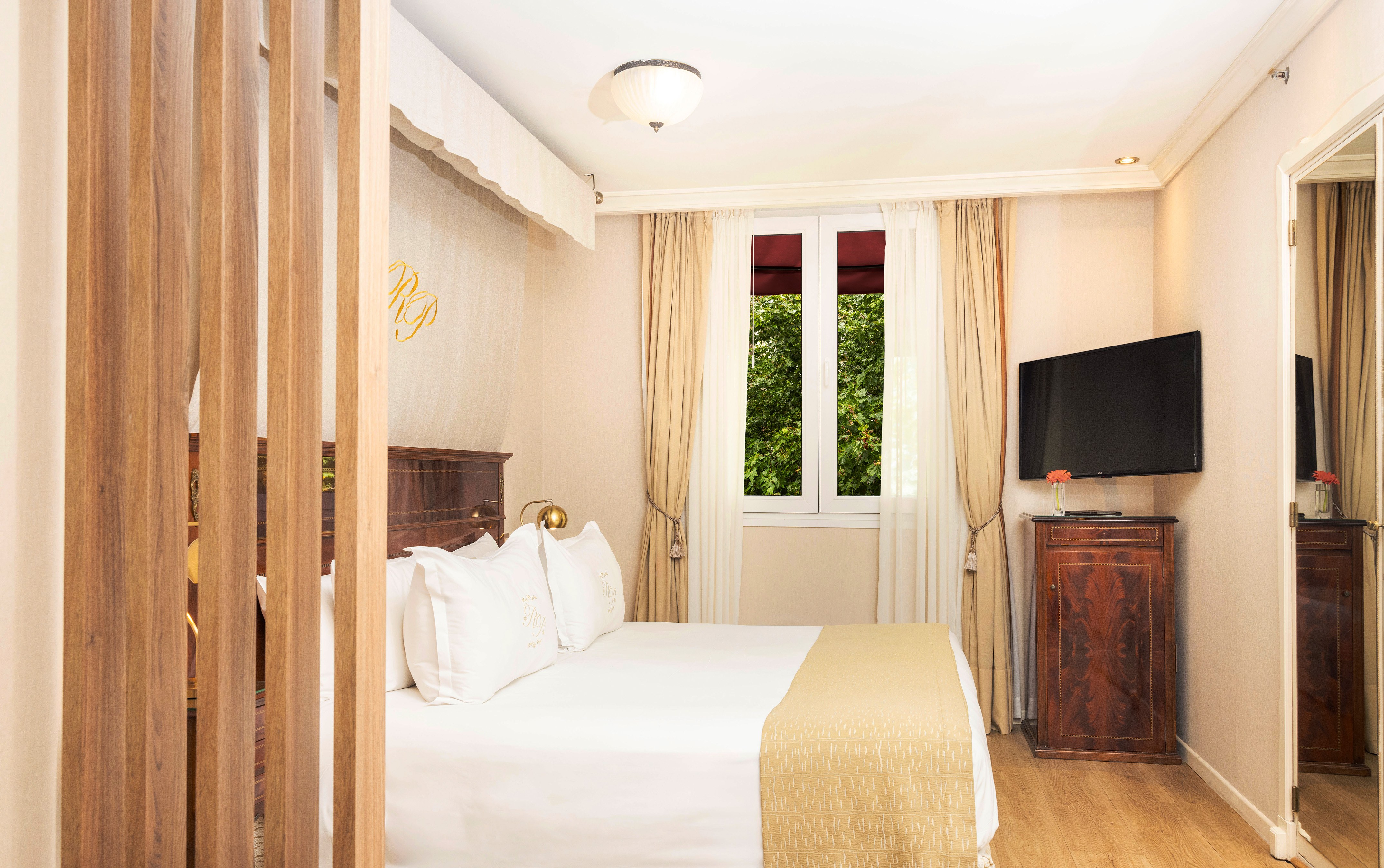 Hotel bedroom with comfortable bed, wooden divider, window view, and mounted TV.