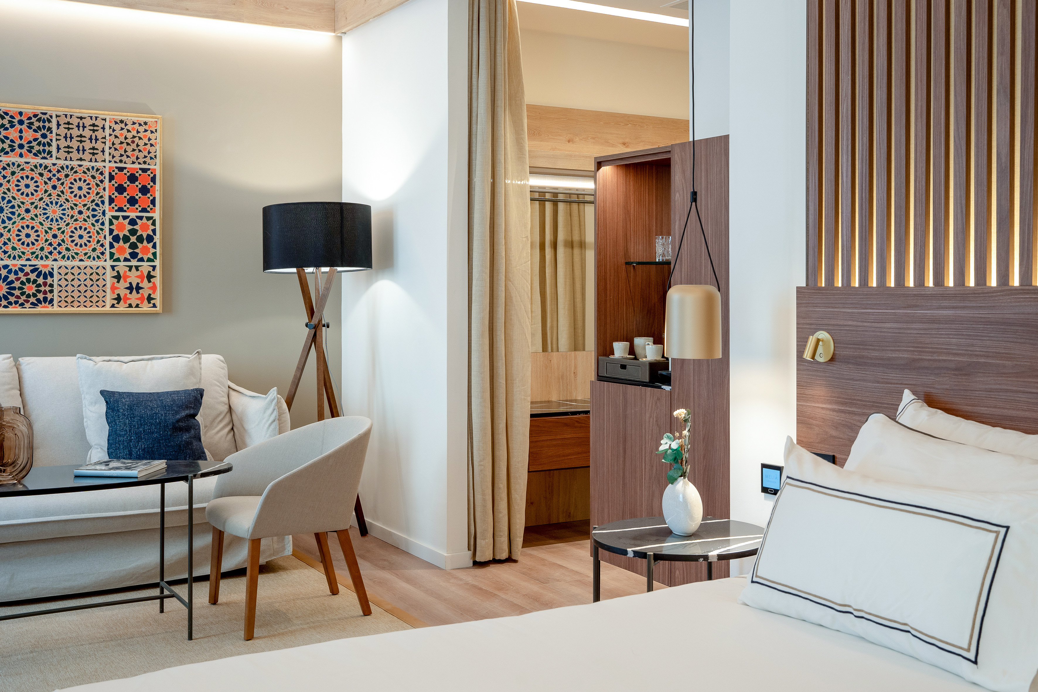 Modern hotel suite with bed, sofa, geometric art, wood details, and warm lighting.