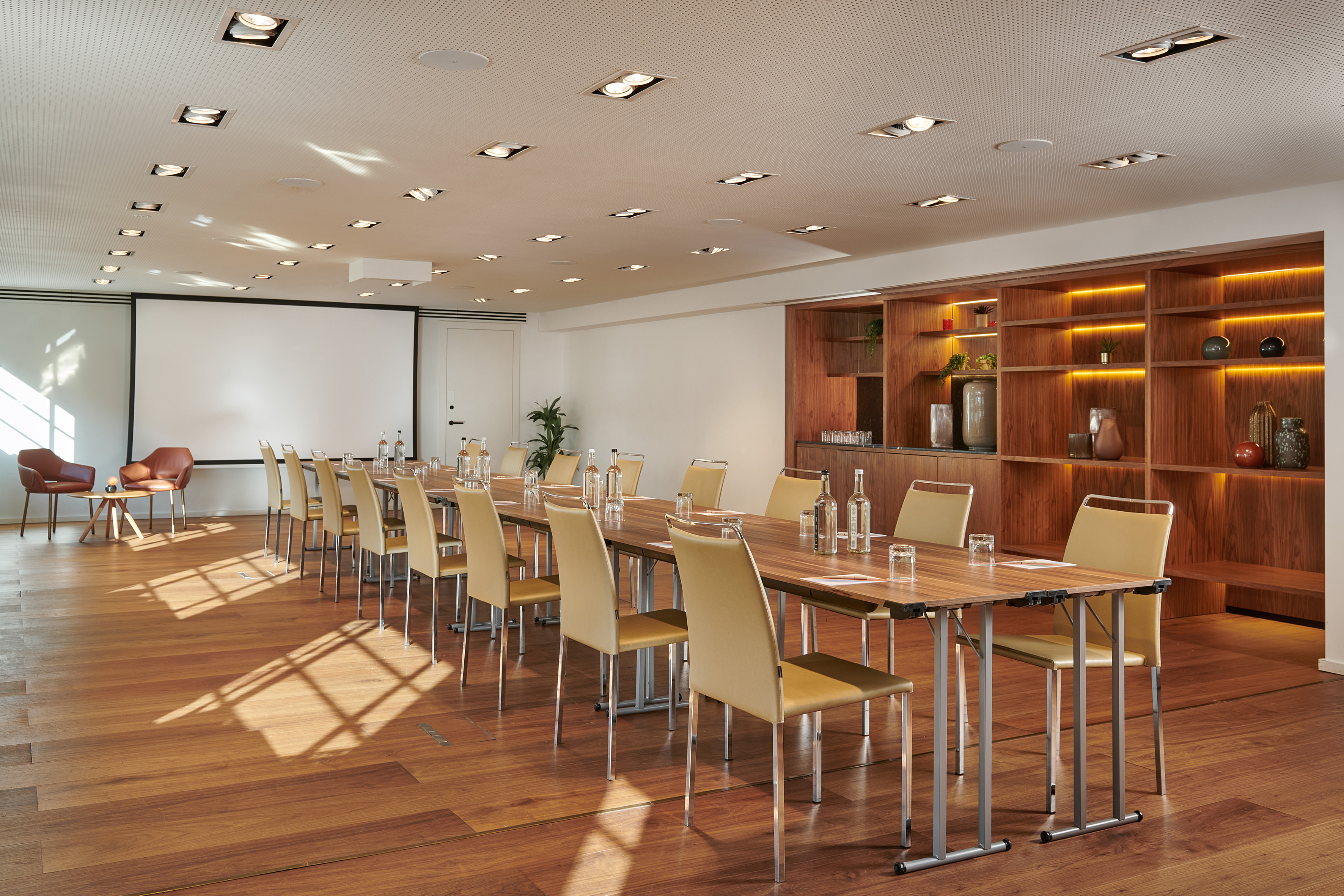 Sunlit modern conference room with long table, projector screen, and wooden shelving.