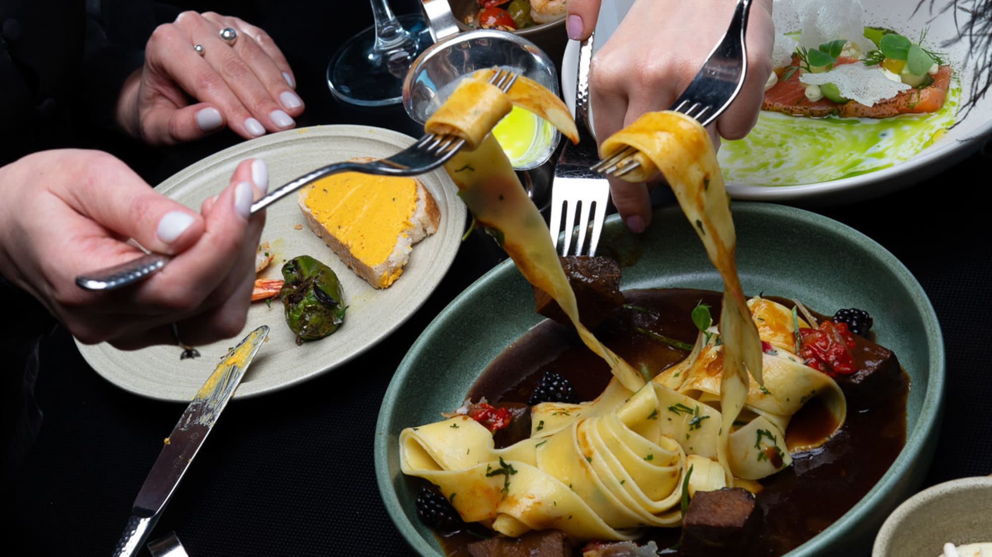 Dining table with hands, gourmet pasta, salmon, peppers, and cocktails.