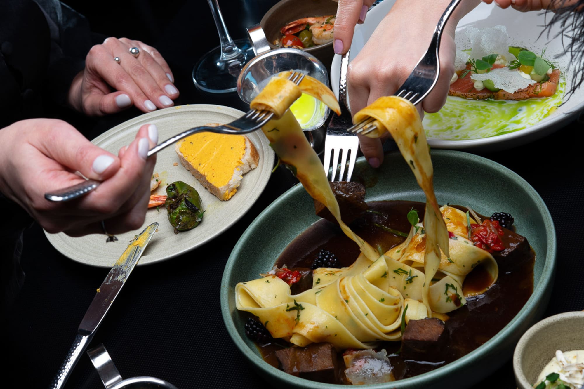 Dining table with hands, gourmet pasta, salmon, peppers, and cocktails.
