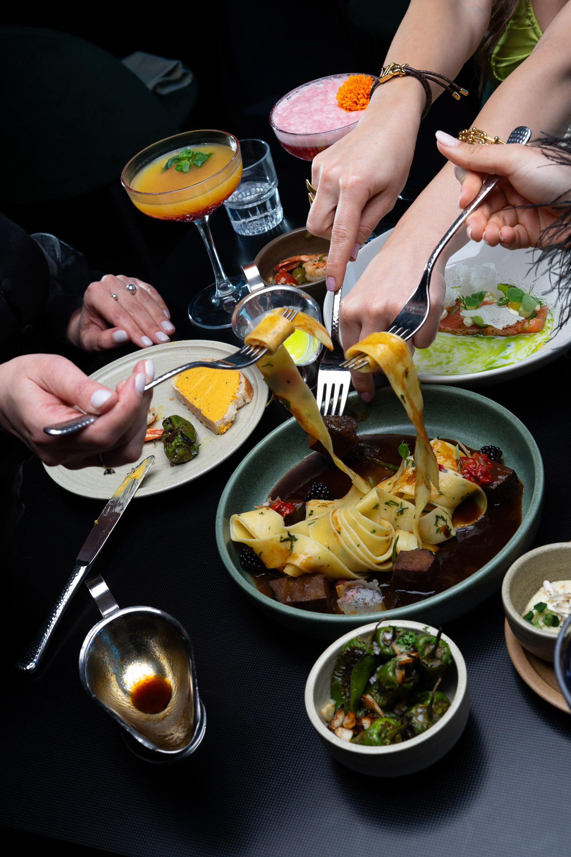 Dining table with hands, gourmet pasta, salmon, peppers, and cocktails.