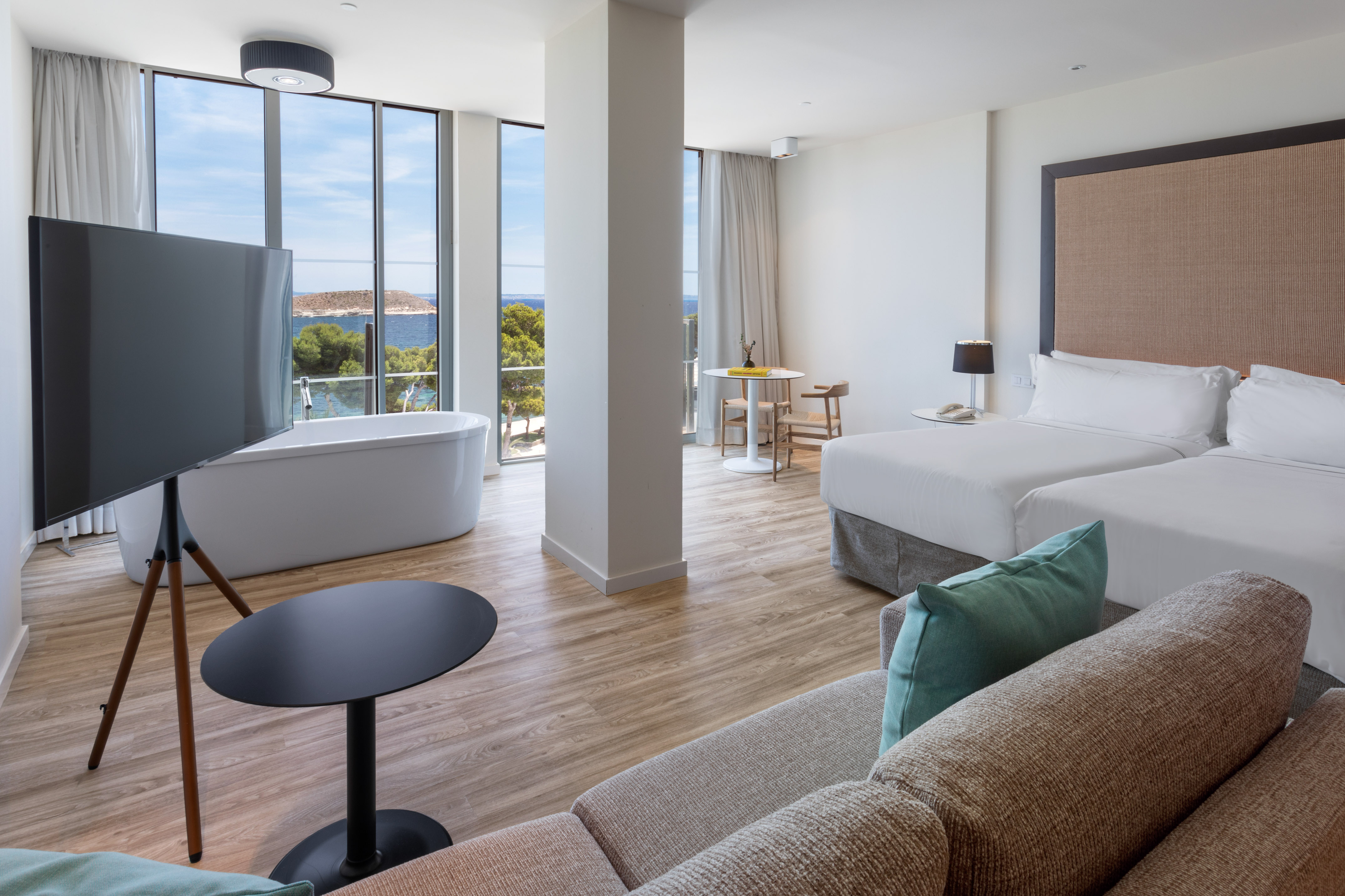 Modern hotel room with sea view, freestanding bathtub, twin beds, and sofa.