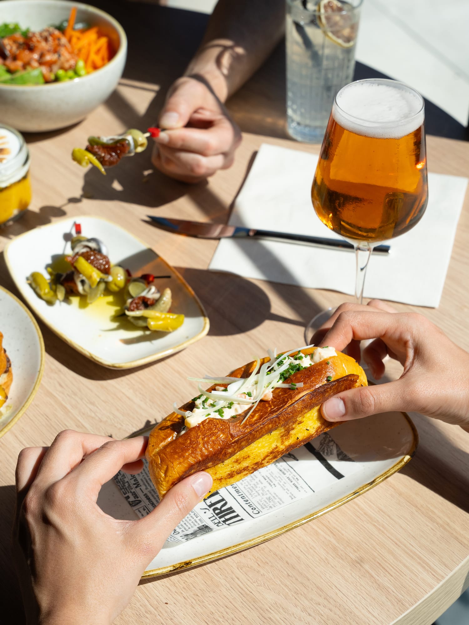 Hands holding a golden lobster roll on brioche, with beer, appetizers, and salad.