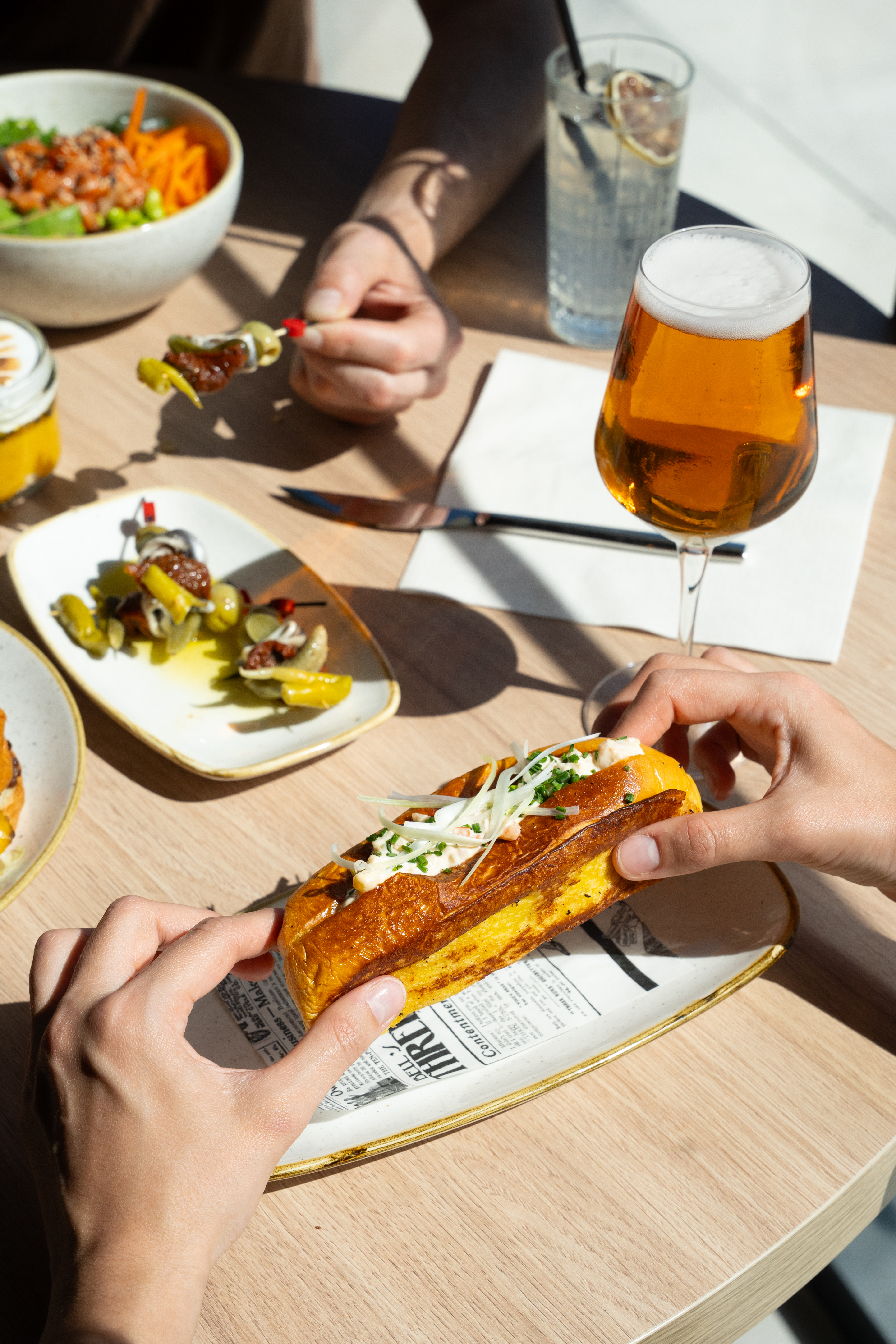 Hands holding a golden lobster roll on brioche, with beer, appetizers, and salad.