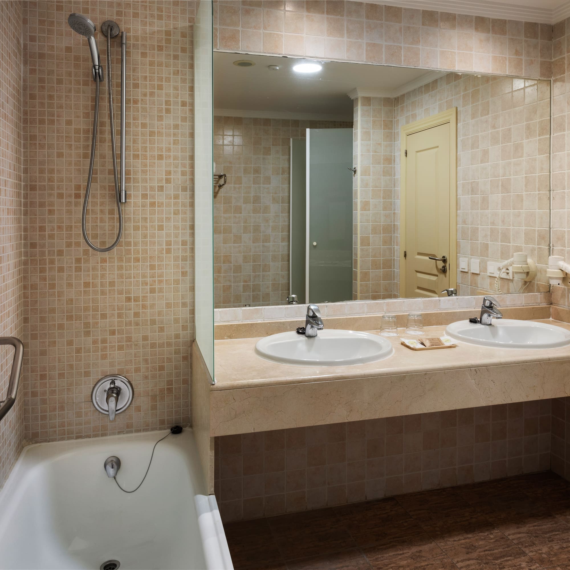 Beige tiled bathroom with bathtub, shower, double vanity, and large mirror.
