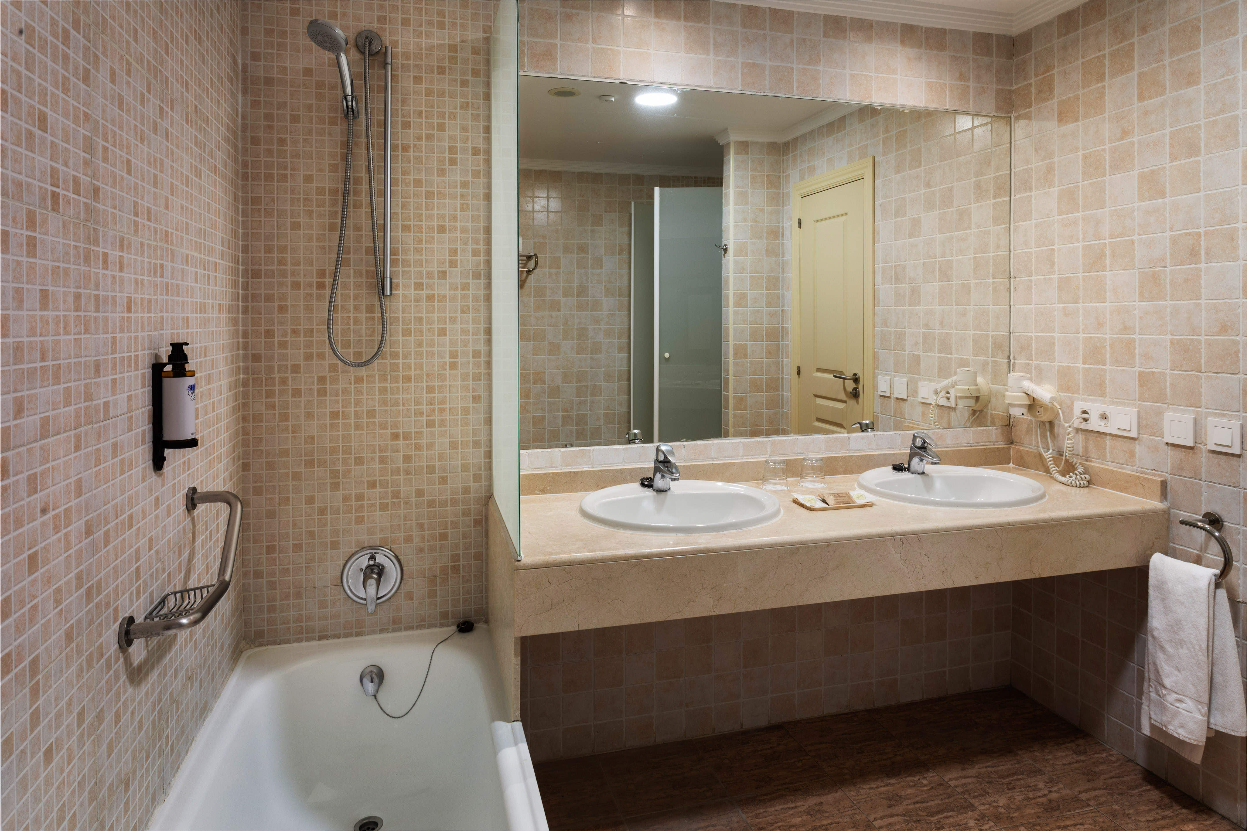 Beige tiled bathroom with bathtub, shower, double vanity, and large mirror.