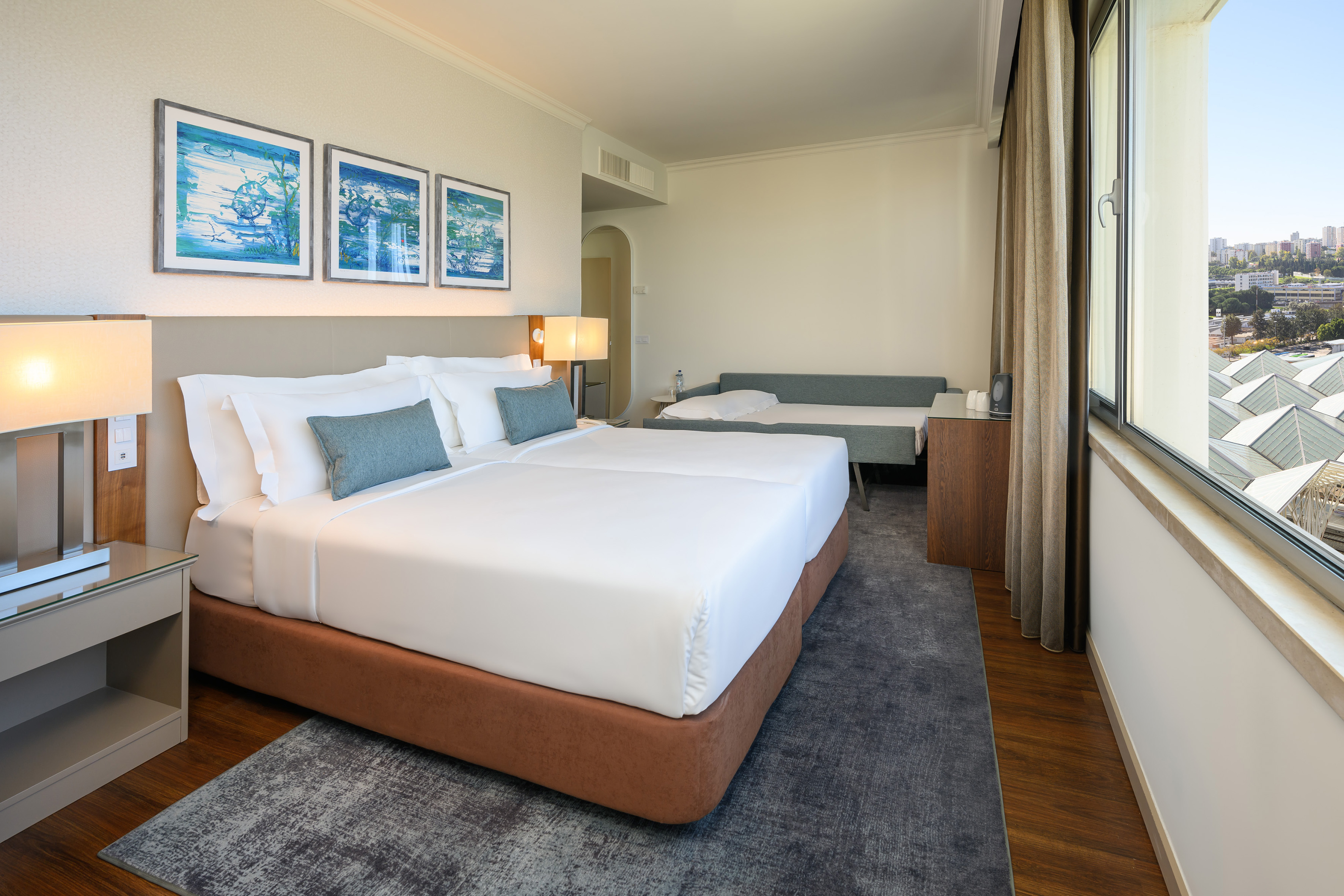 Modern hotel room with twin beds, sofa bed, city view, and blue art.