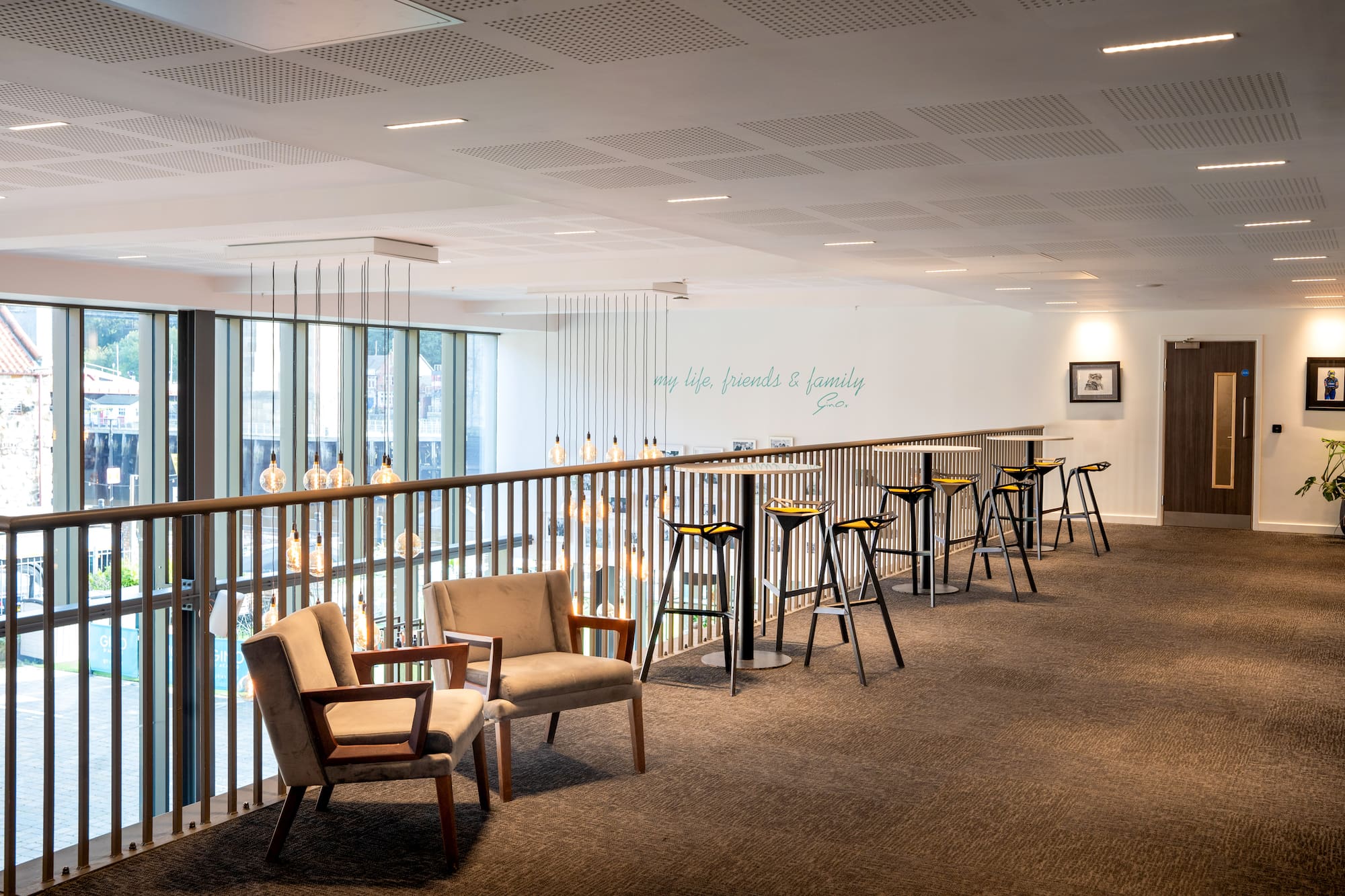 Modern office lounge with armchairs, bar stools, window view, and hanging lights.