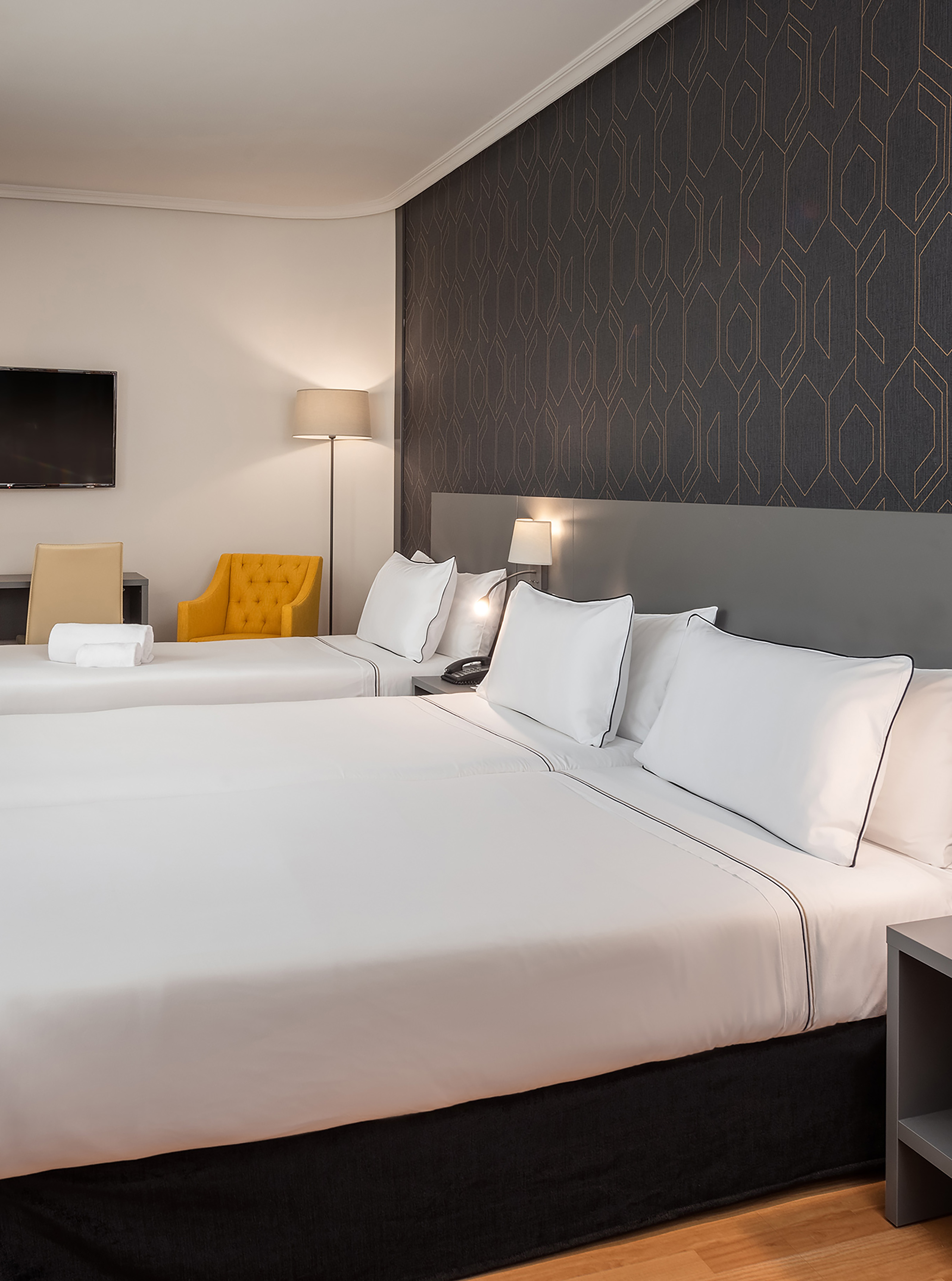 Modern hotel room with two twin beds, yellow armchair, and geometric wallpaper.