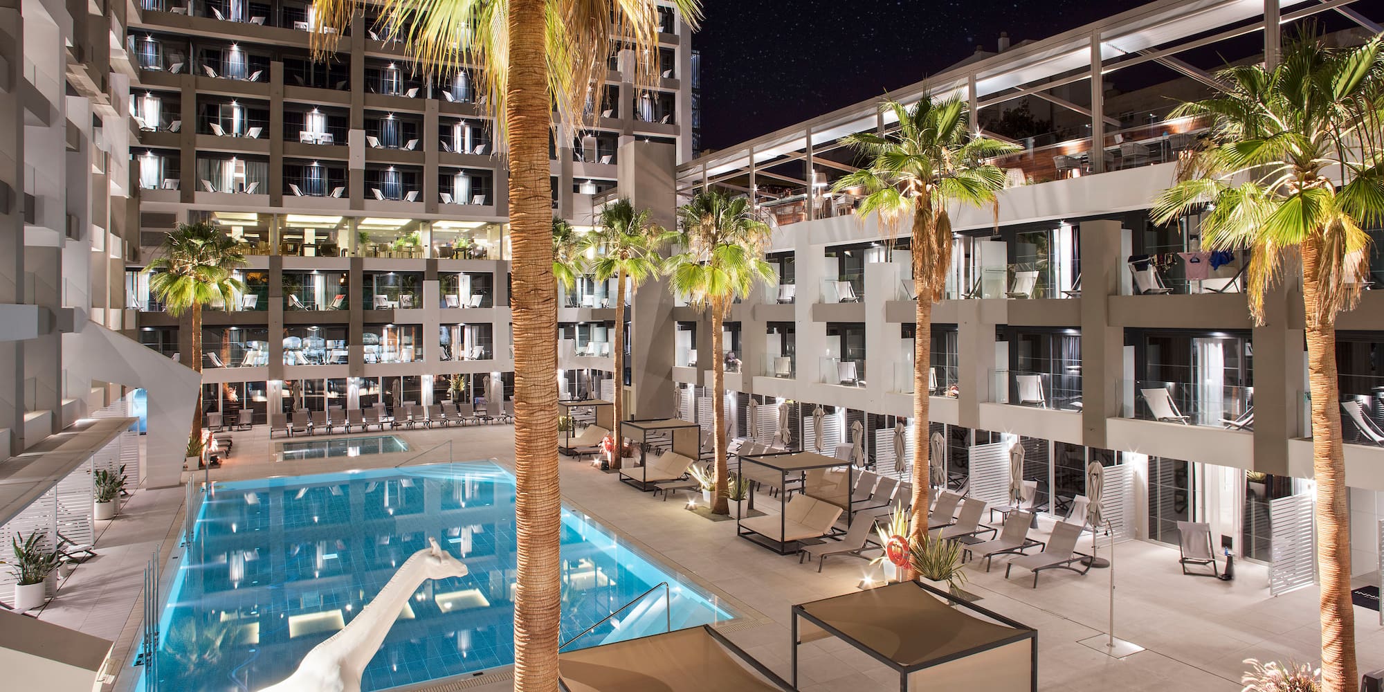 Hotel moderno de noche: piscina, palmeras, terraza, jirafa blanca bajo estrellas.