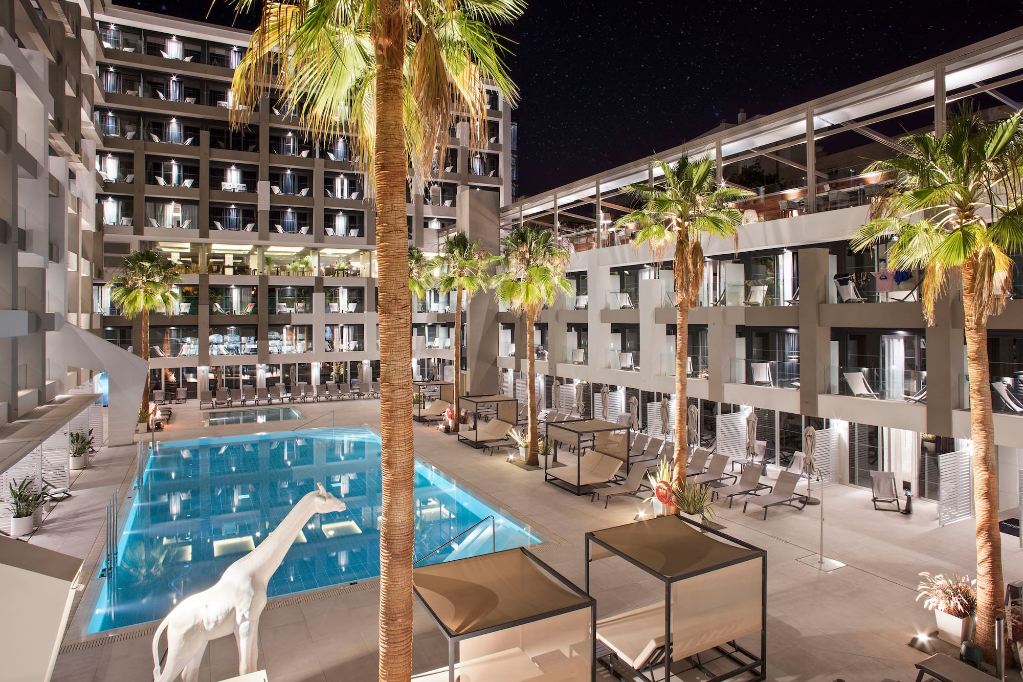 Nighttime luxury hotel resort with illuminated pool, palm trees, and cabanas.