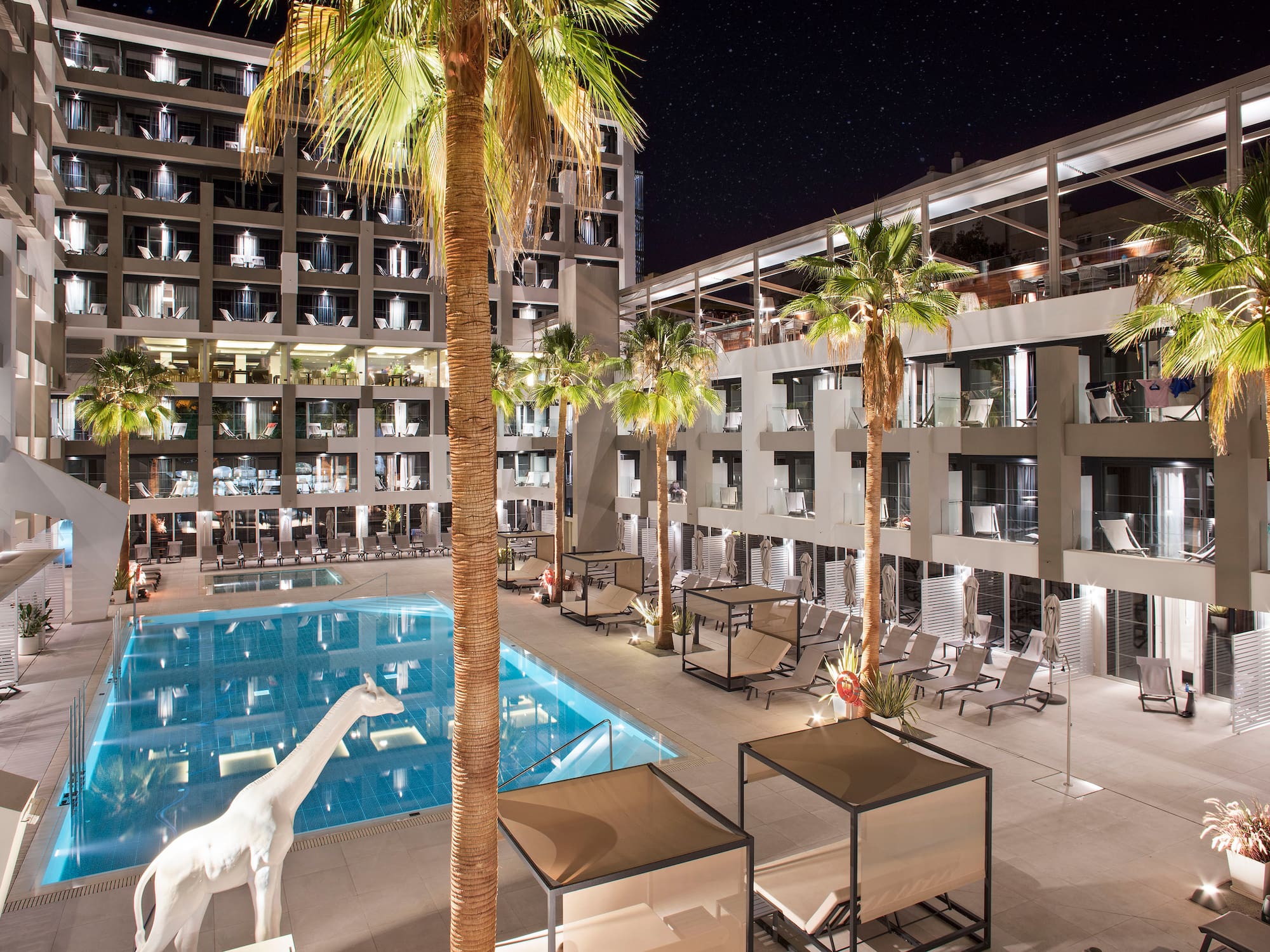 Hotel moderno de noche: piscina, palmeras, terraza, jirafa blanca bajo estrellas.