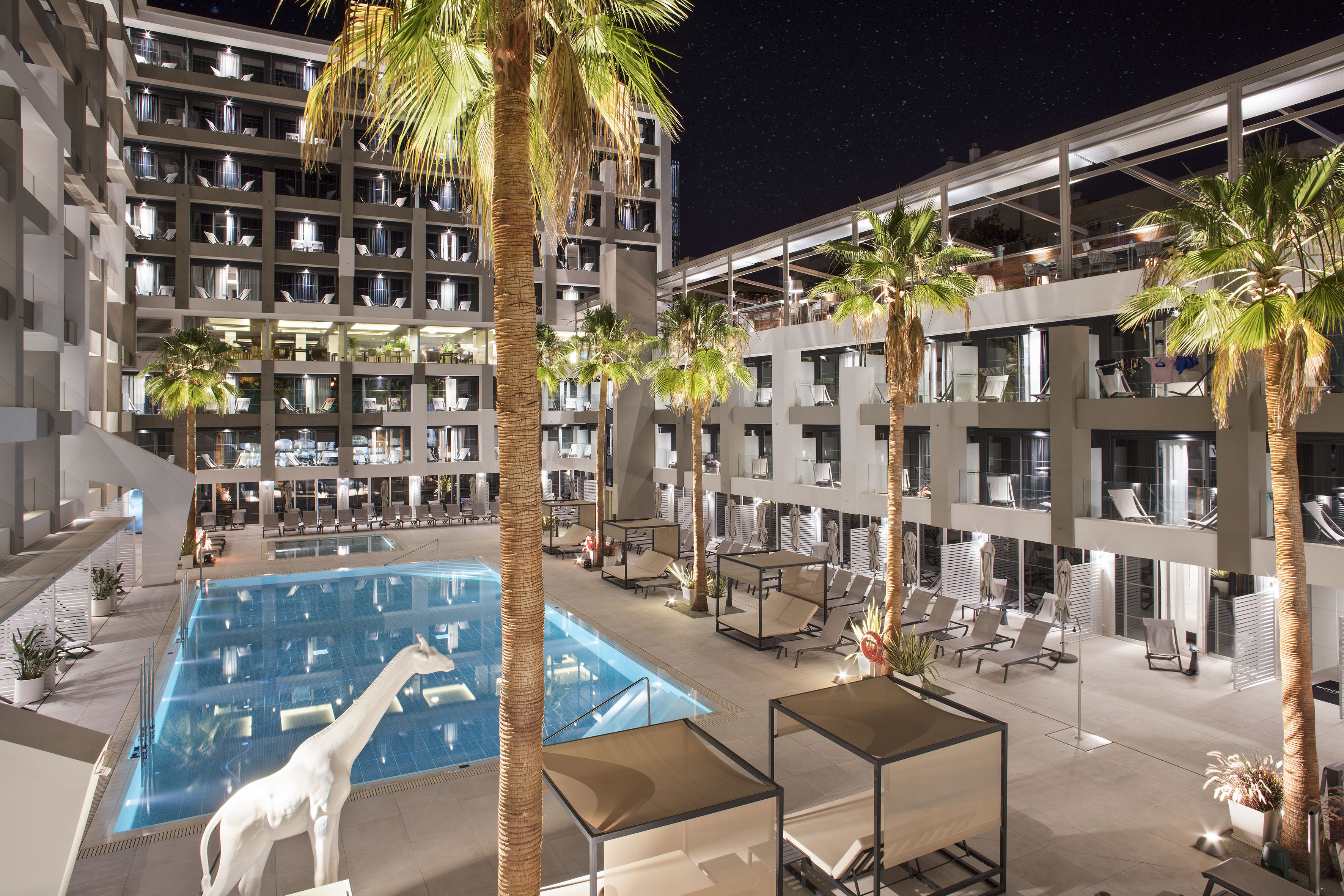 Hotel moderno de noche: piscina, palmeras, terraza, jirafa blanca bajo estrellas.