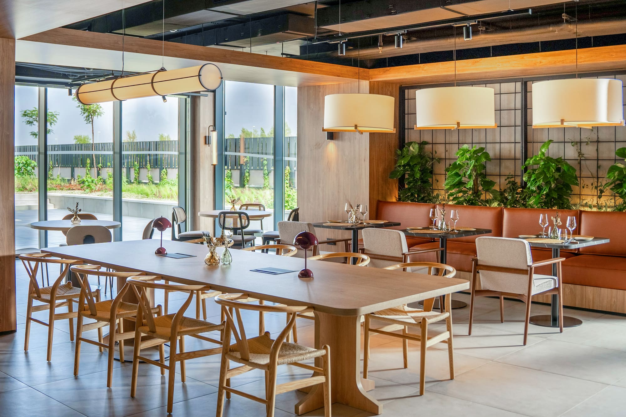 Stylish restaurant with long wooden table, wishbone chairs, booth seating, and greenery.