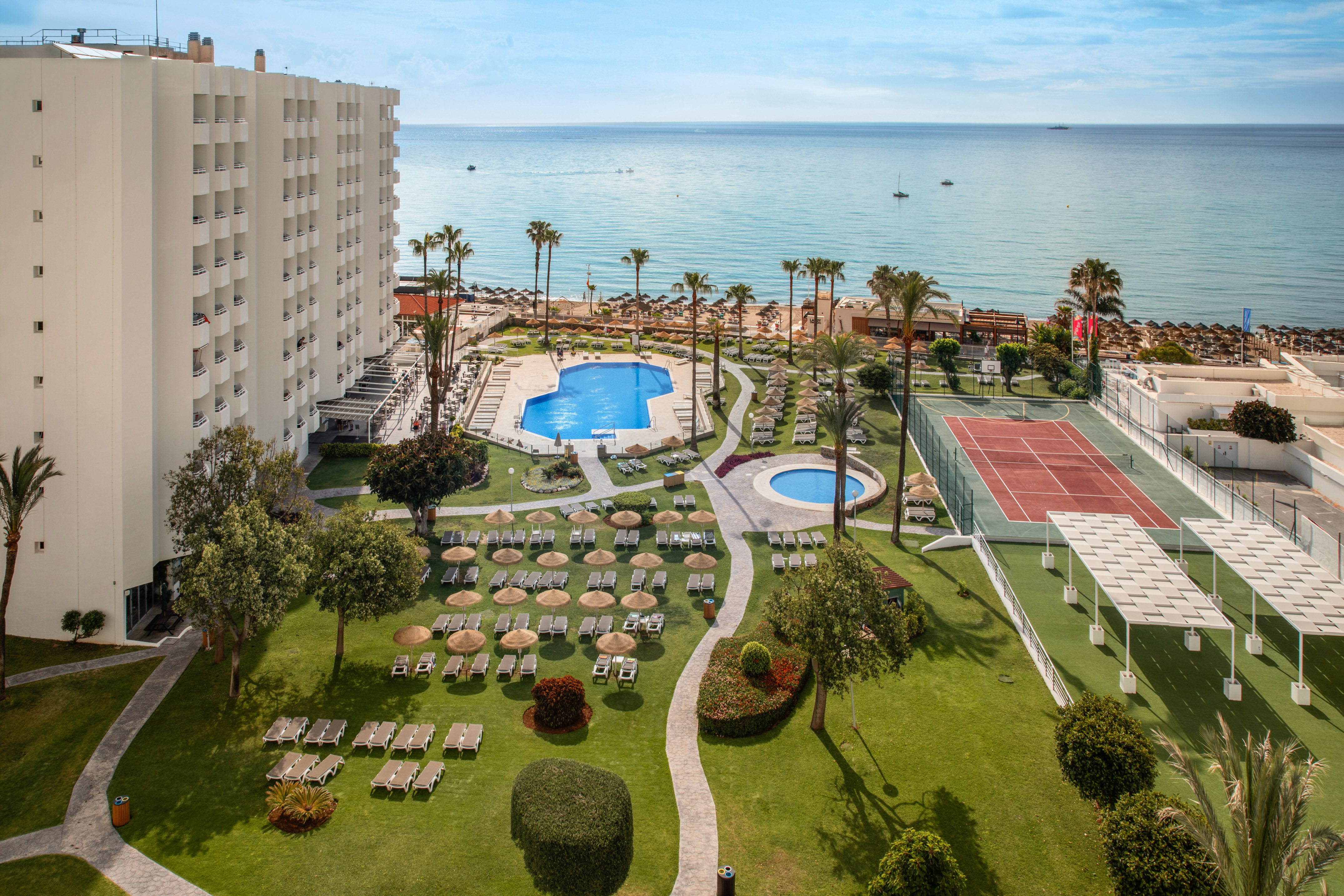Beachfront hotel with swimming pools, tennis court, palm trees, and ocean.