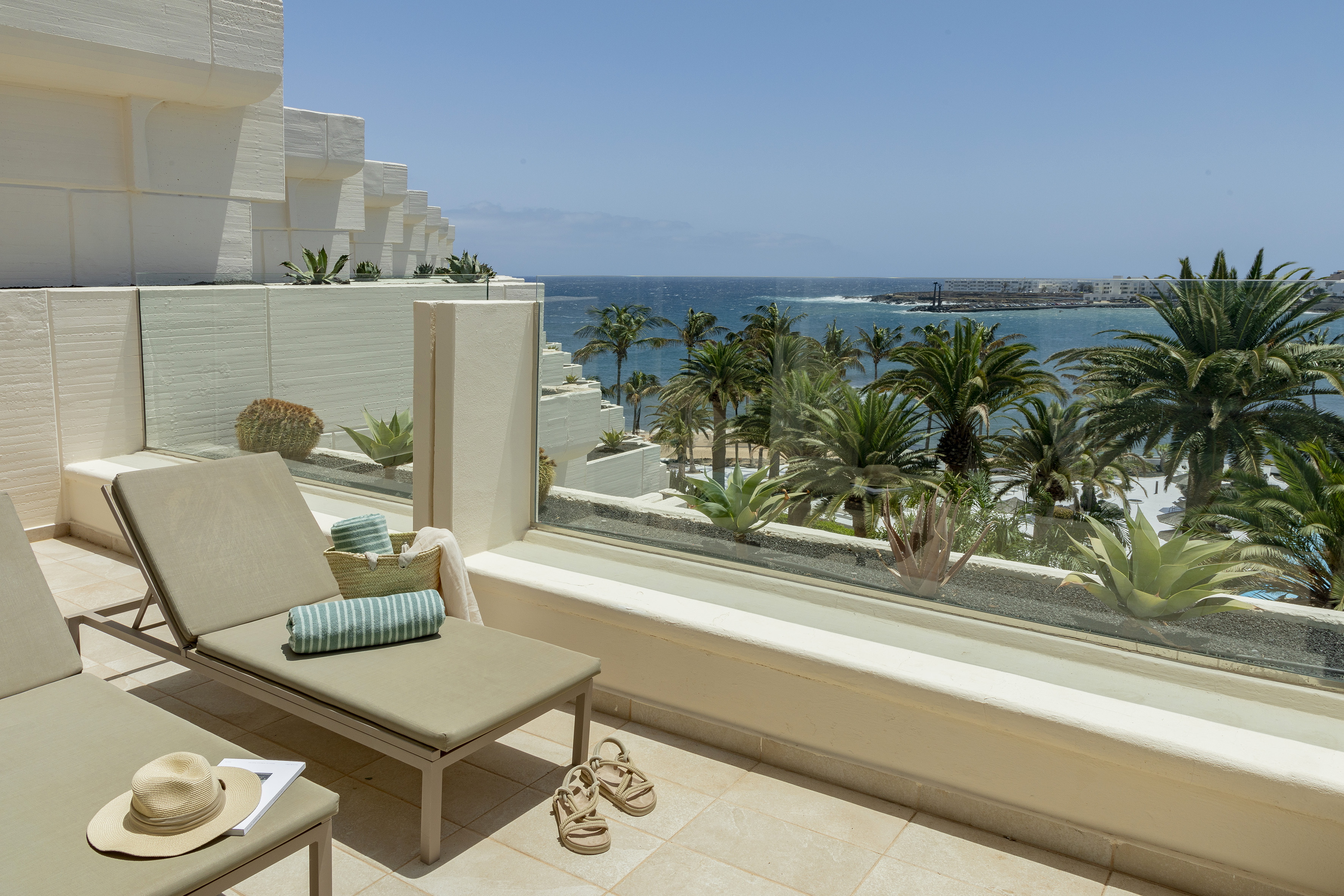 Sunny hotel balcony with ocean view, palm trees, and loungers.