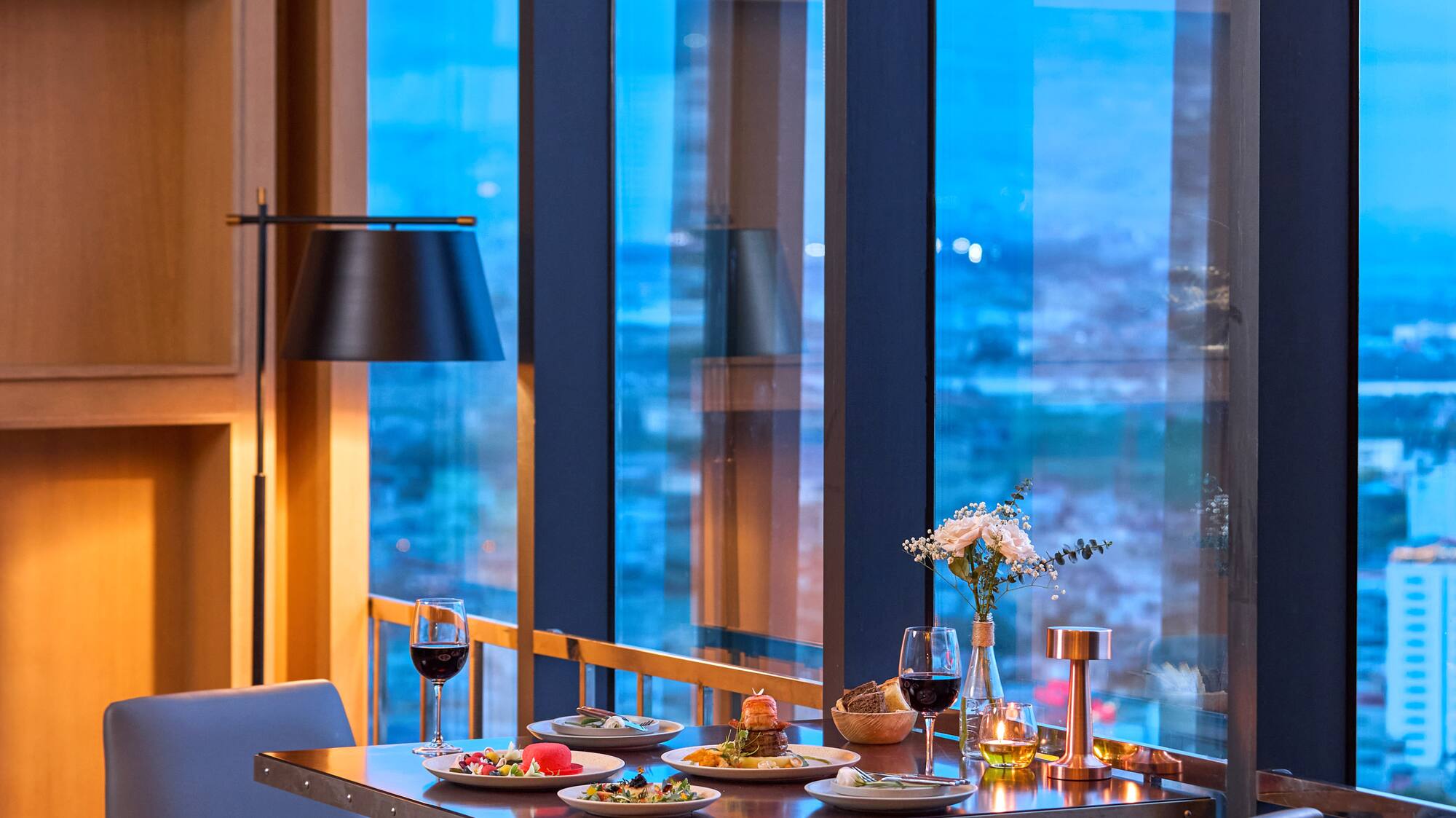 Romantic dinner table with wine, modern chairs, city skyline view at dusk.